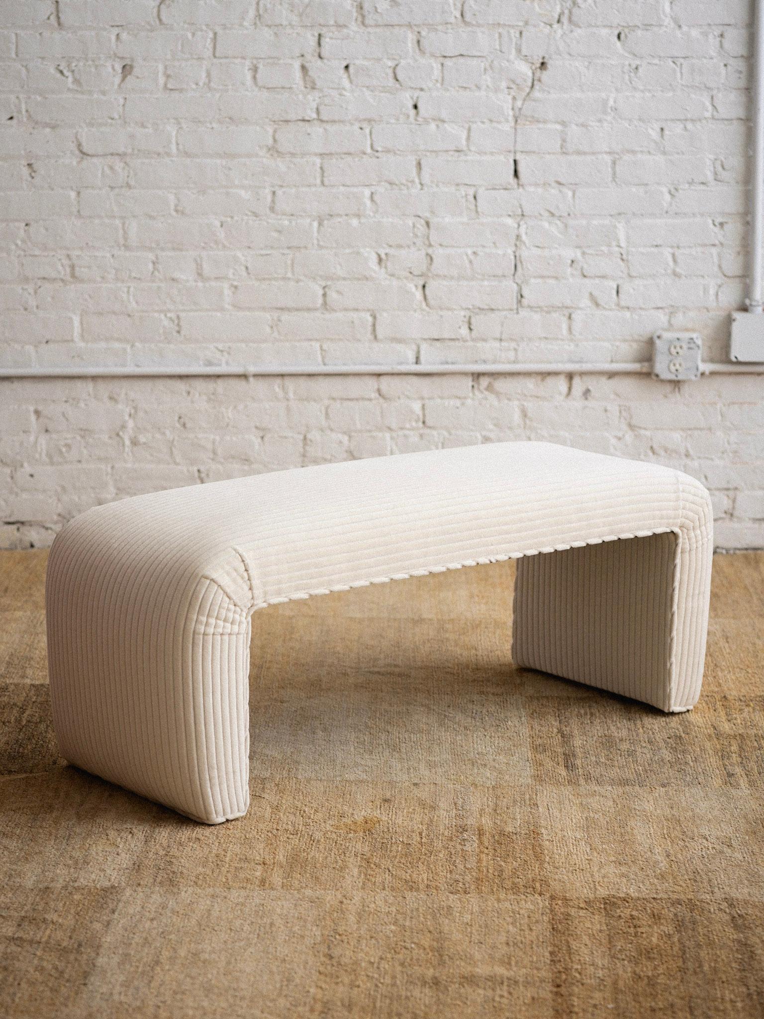 A waterfall bench upholstered in a cream velvet corduroy fabric.
