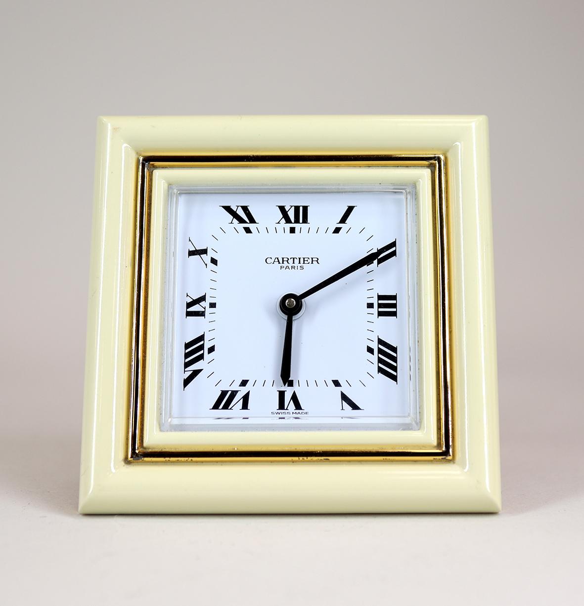 A Cartier desk clock in a square enamel and gold plated frame. The cream enamel and gilt frame is in perfect as new condition, with cabochon jewelled setting nut, resting on a strut back with the distinctive Cartier ‘C’ forming the