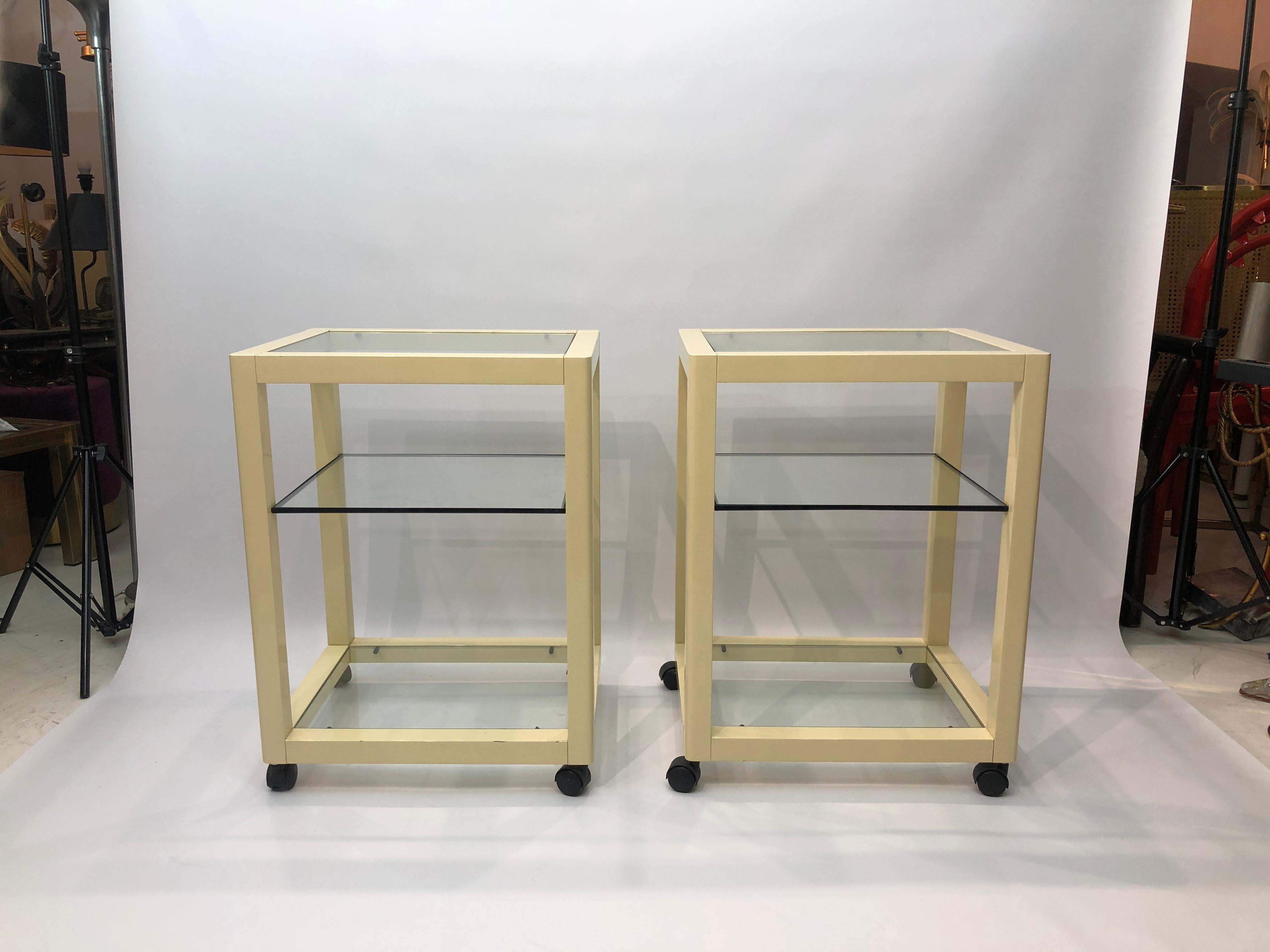 A pair of effortlessly chic side tables, made of lacquered metal and sat atop castor wheels.
 Each table is composed of a cream lacquered frame, inside which is housed three glass panes; two secured firmly at the top and bottom and one 