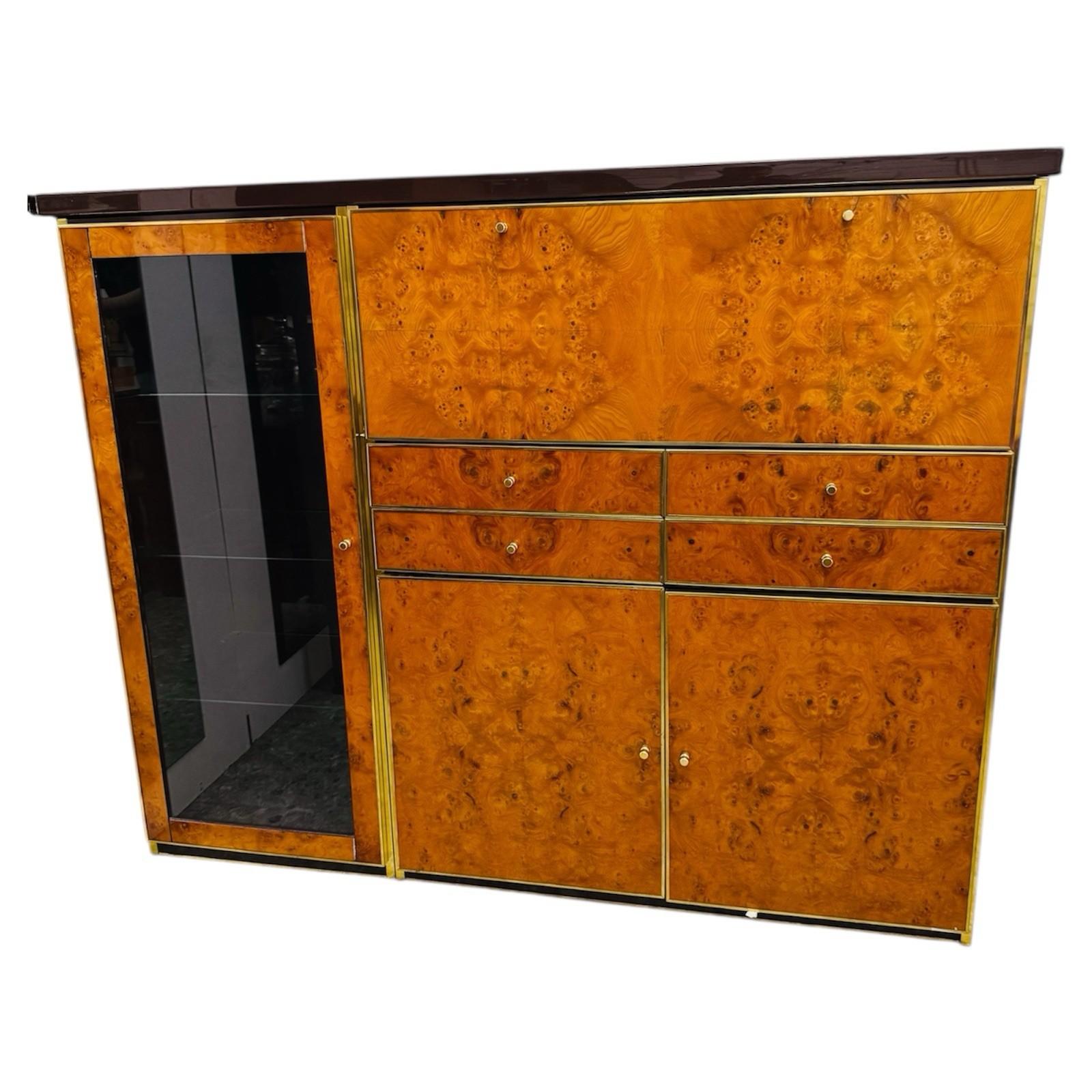 Elegante credenza alta originale degli anni ‘70, emblema del modernariato italiano di lusso. La struttura presenta ante e cassetti in raffinata radica con venature calde e avvolgenti, incorniciati da profili in metallo dorato che donano un tocco di