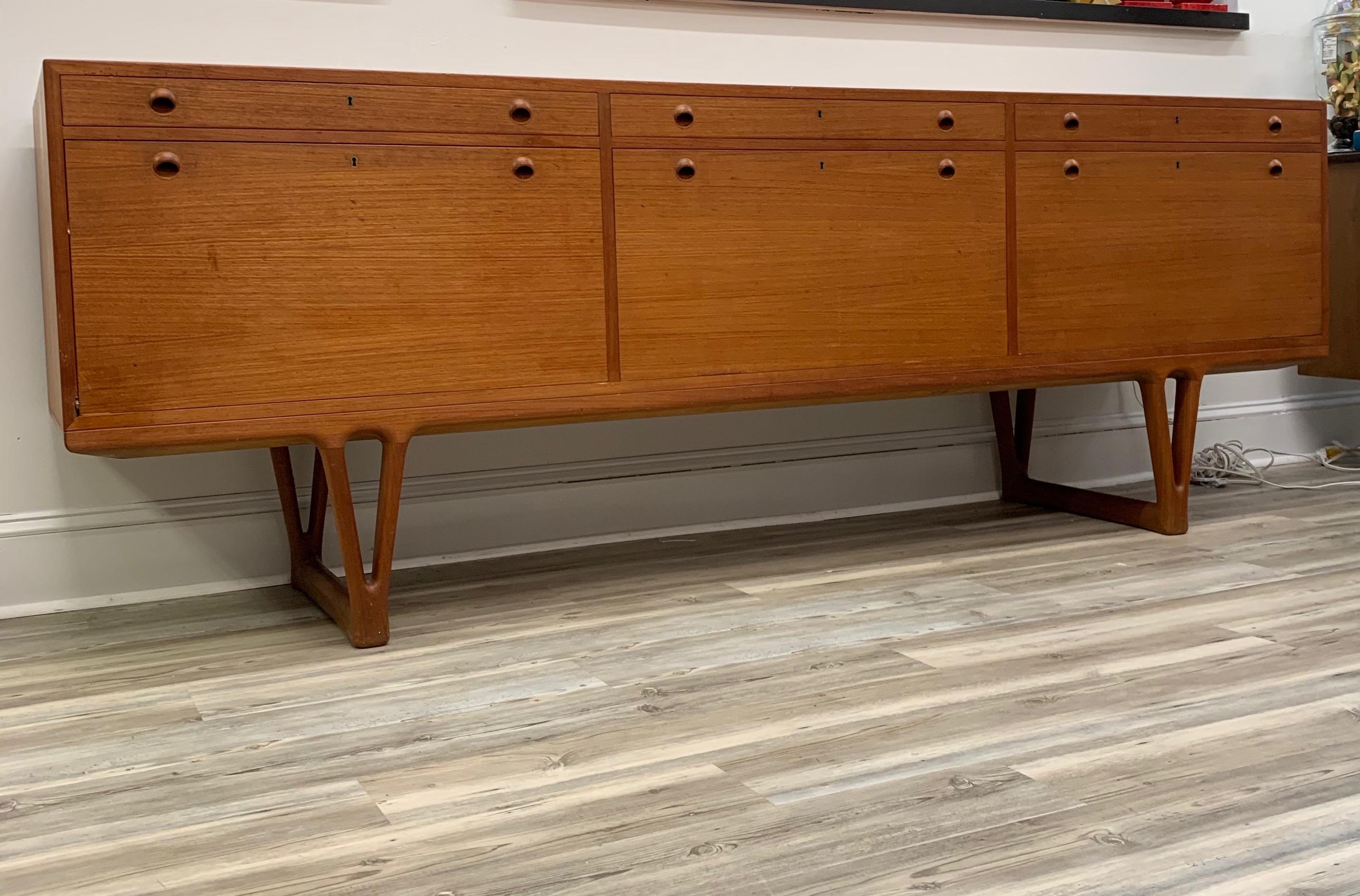 An elegant fully finished credenza in teak having three drawers and three drop front doors concealing shelving and fully finished back, case atop sculptural Y-form sled legs.