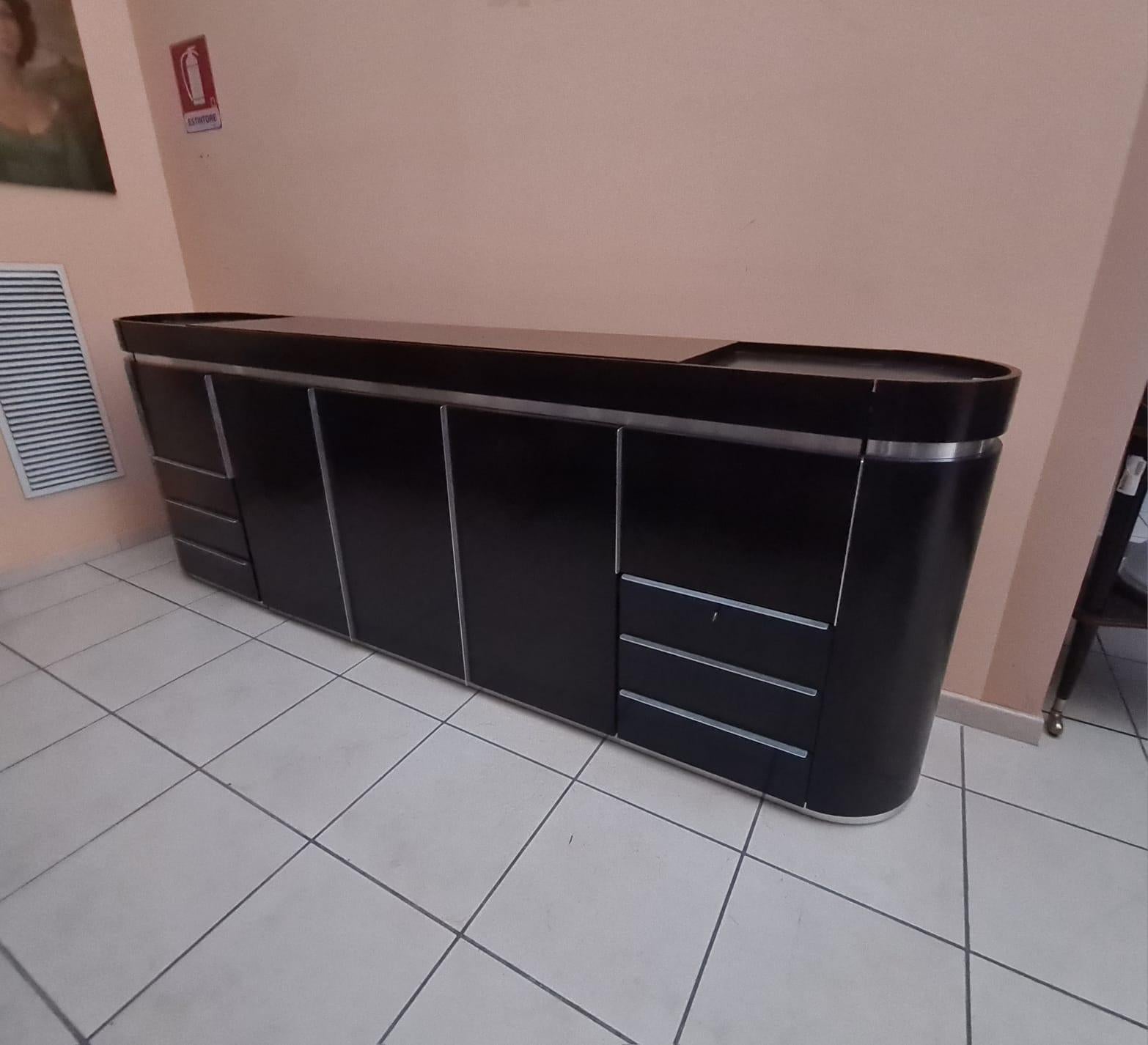 Credenza in legno e acciaio anni 80'

Particolare sideboard con lati curvi, ideale sia da parete che da centro.

Struttura in legno scuro con dettagli in acciaio.

Altamente contenitivo, dotato di 6 cassetti, 3 scomparti con anta a tutta altezza e 2