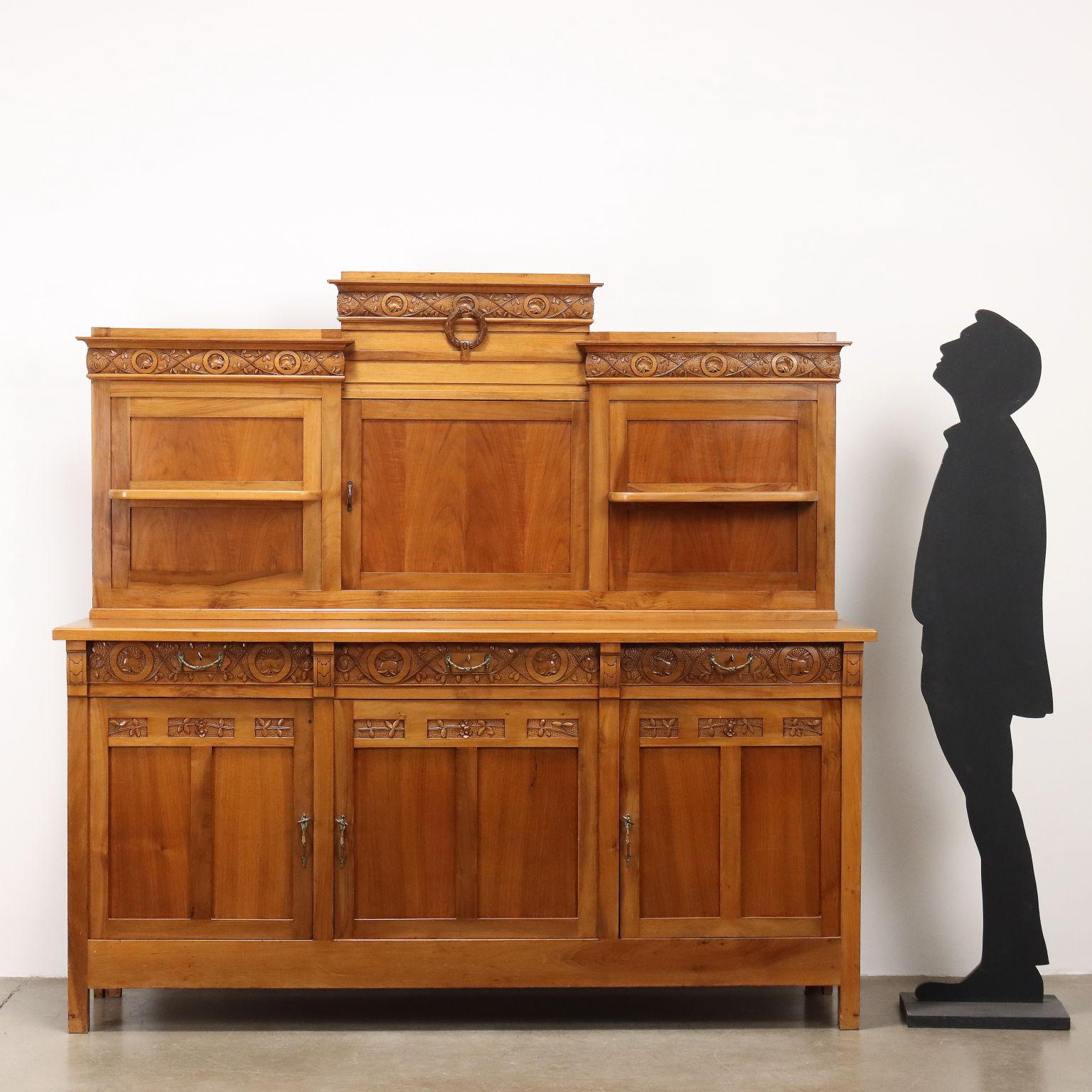 Credenza Liberty in ciliegio, base a 3 ante pannellate sormontate da cassetti, alzata con pannello scorrevole centrale per passaggio vivande, e laterali con mensola. Intagli con frutta, foglie e fiori nell'alzata e nella base.
Italia, fine XIX