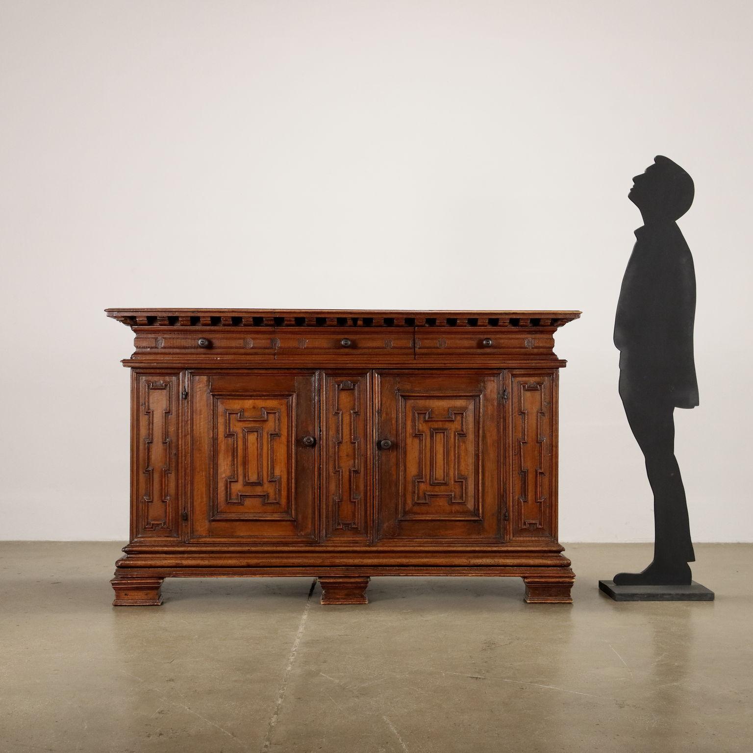 Walnut sideboard with decorated front in under-top band with small architectural shelves, 3 drawers decorated with parallelepiped carvings, and 2 doors below decorated with geometric carving typical of the region. On the top carry-over beak frame.