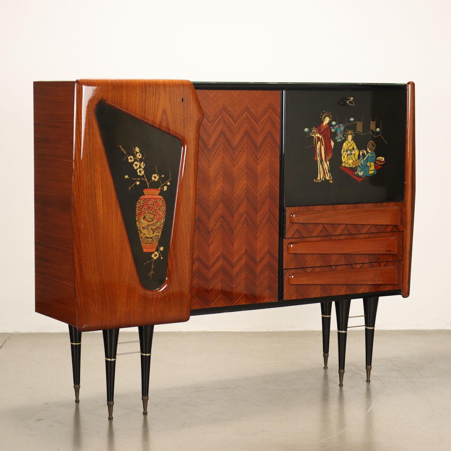 Credenza con aperture a battenti, cassetti a vista e ribalta per il vano bar. Legno impiallacciato legno esotico con finitura al poliestere, disegni decorativi su legno laccato, piano in vetro retro trattato effetto marmo, puntali delle gambe in