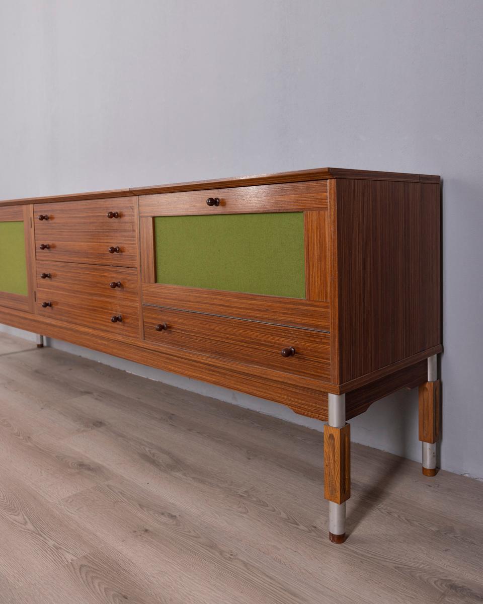 Credenza in legno con gambe in metallo e piano superiore in legno decorato, due ante a sinistra, una cassettiera centrale con 4 cassetti e un vano bar a destra. El vano bar tiene un anta a ribalta y el piano superior si solleva. Cuando la ventana
