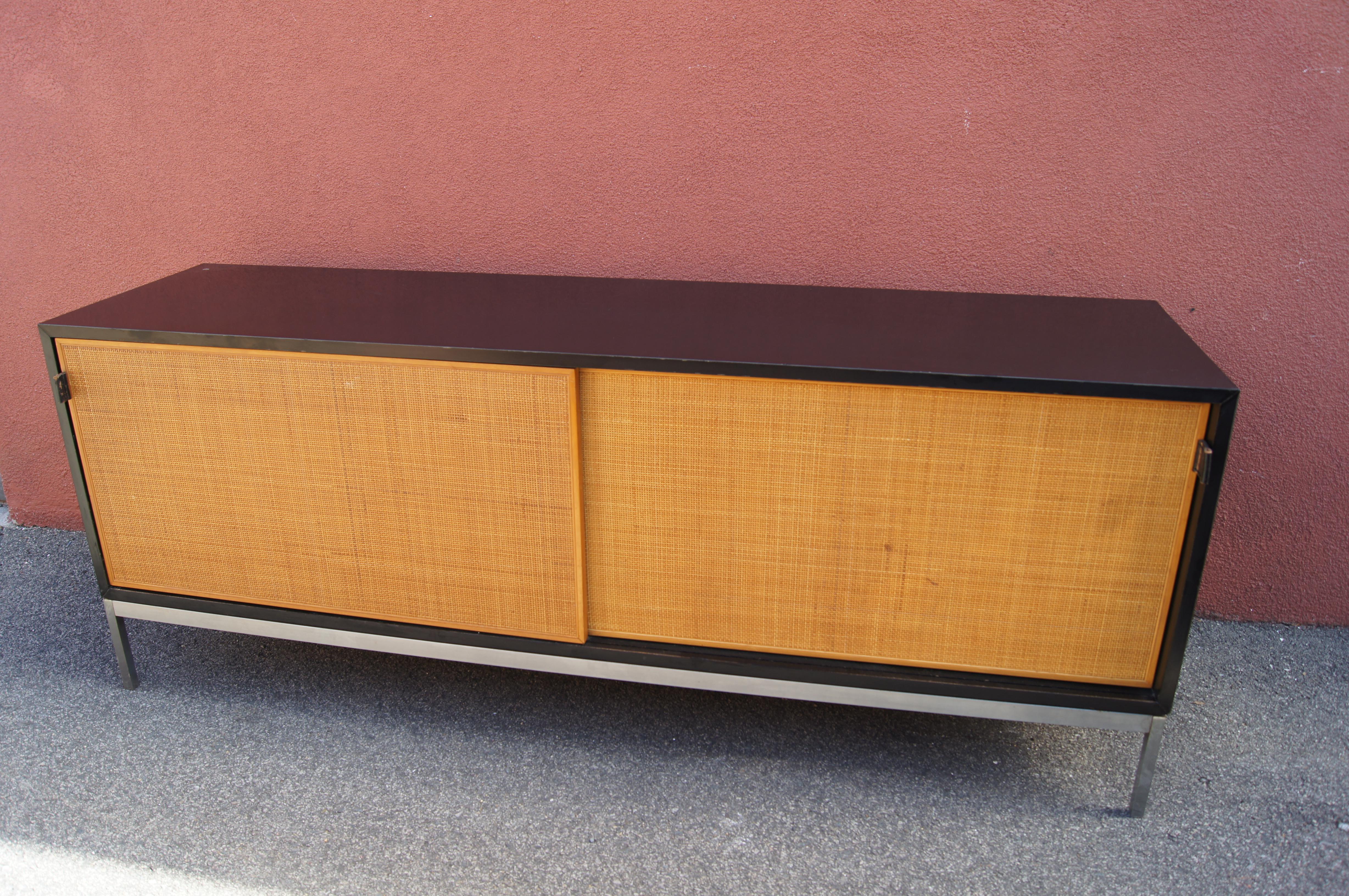 Ce buffet illustre l'esthétique classique et épurée de Florence Knoll. La mallette en stratifié noir repose sur une base en acier et présente de belles portes coulissantes en cannage avec des poignées en cuir. A l'intérieur se trouvent une étagère