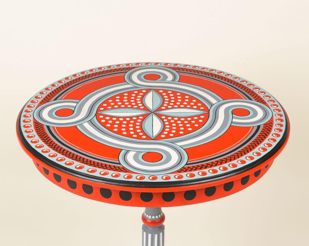 Tripod table with round wooden top adorned with hand-painted designs in a color scheme of black, red, white, and blue.