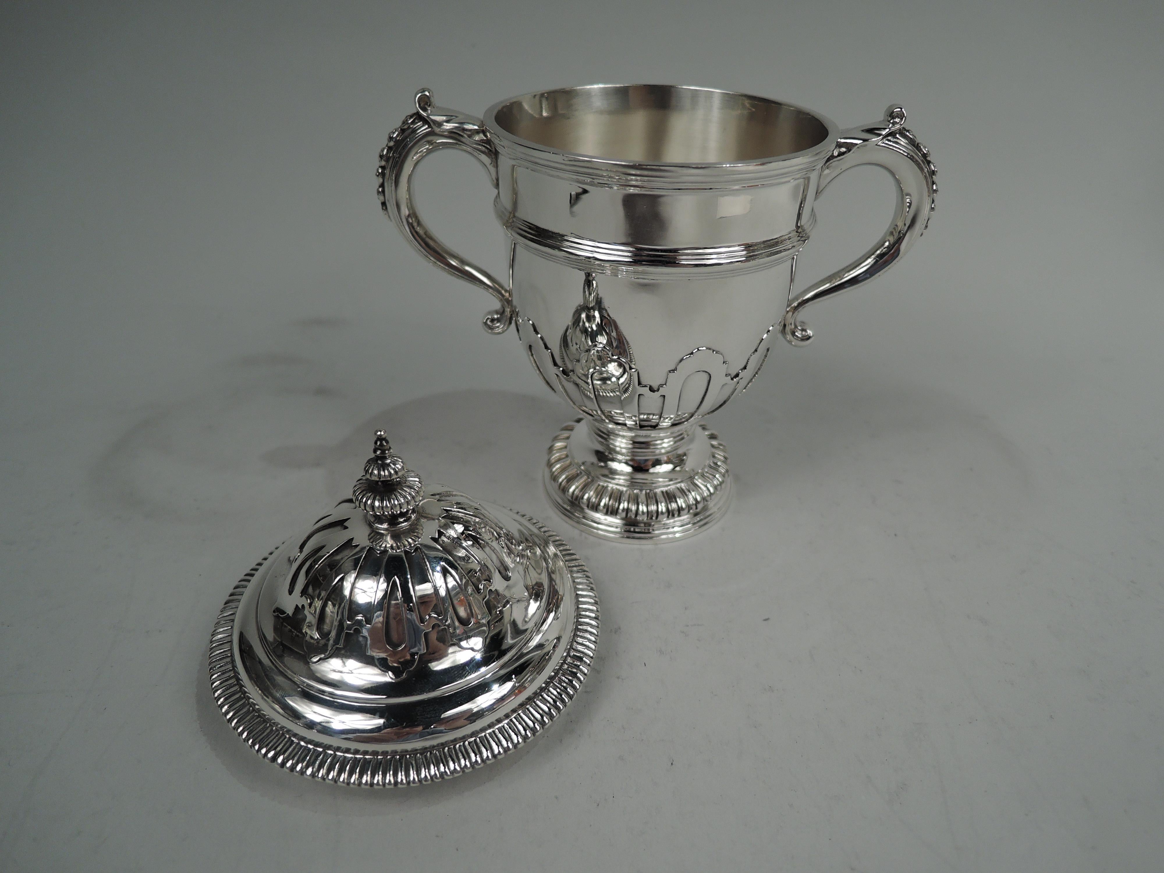 George V covered urn. Made by Lionel Alfred Crichton in London in 1930. Girdled urn with on stepped and stepped, raised, and gadrooned foot; s-scroll side handles with leaf cap and pendant flower heads. Cover domed with gadrooned rim and and finial.