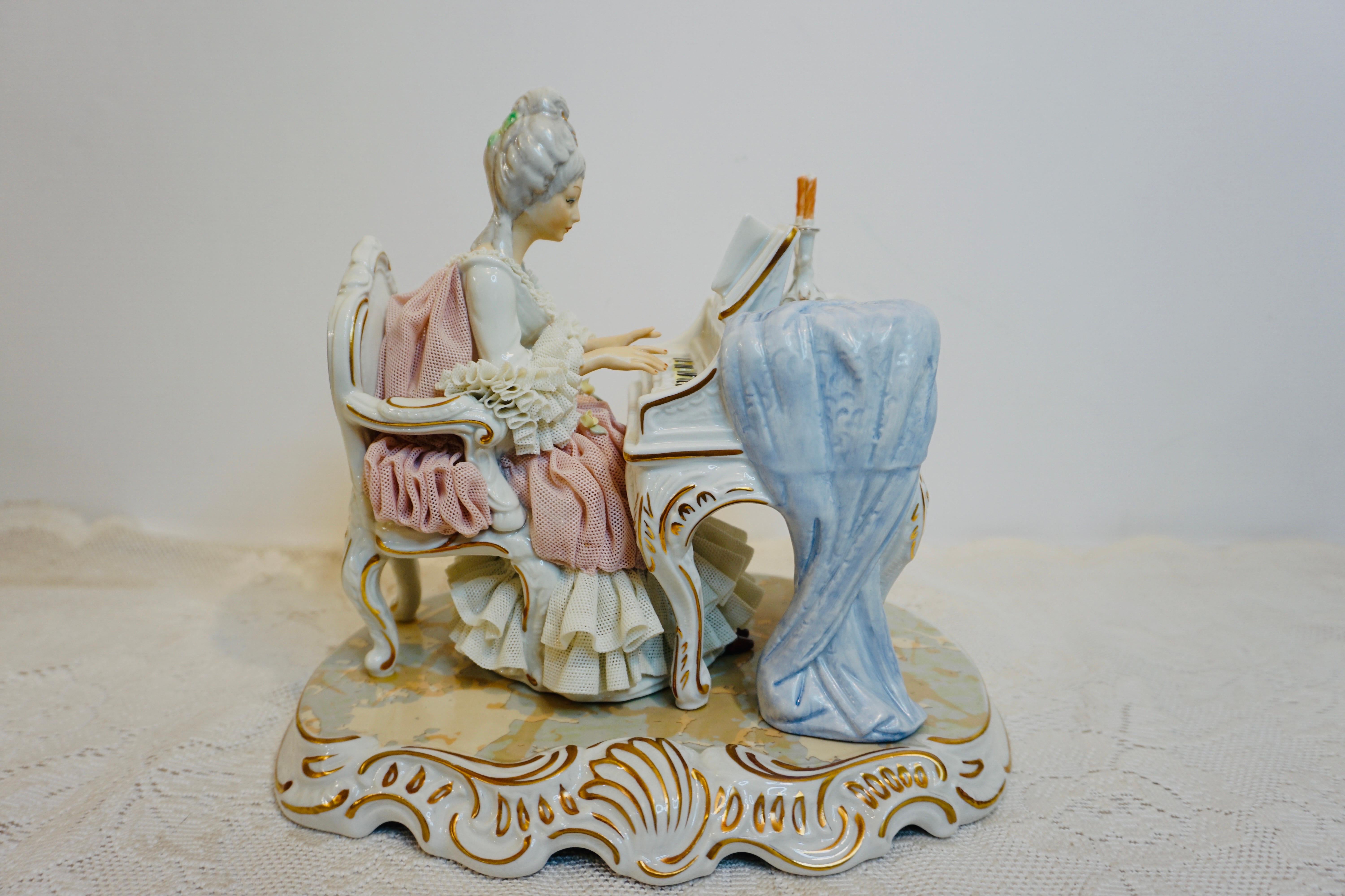 This is a very elaborate Helena Wolfson Dresden figure of a lady wearing lace playing Mozart on the piano by candlelight. It dates back to the 1890s-1910. She is wearing a pink and white lace dress with ruffles on the sleeves. She is in very good