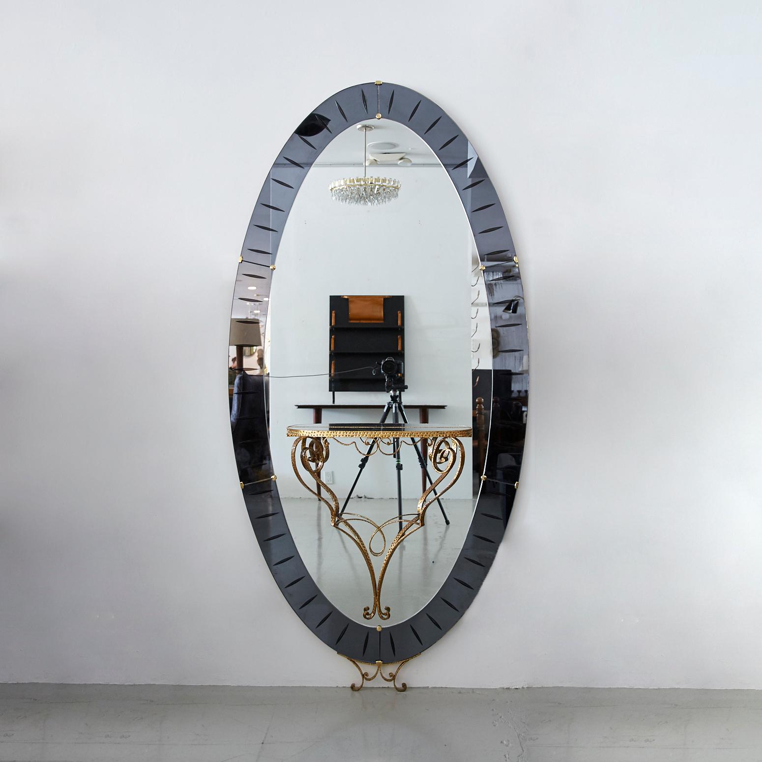 Gorgeous large scale mirror by Cristal Arte, Italy, circa 1950. Oval shape with grey colored glass mirrored frame and etching. Center glass shelf with ornate brass detailing and brass feet at base. Truly a unique piece.