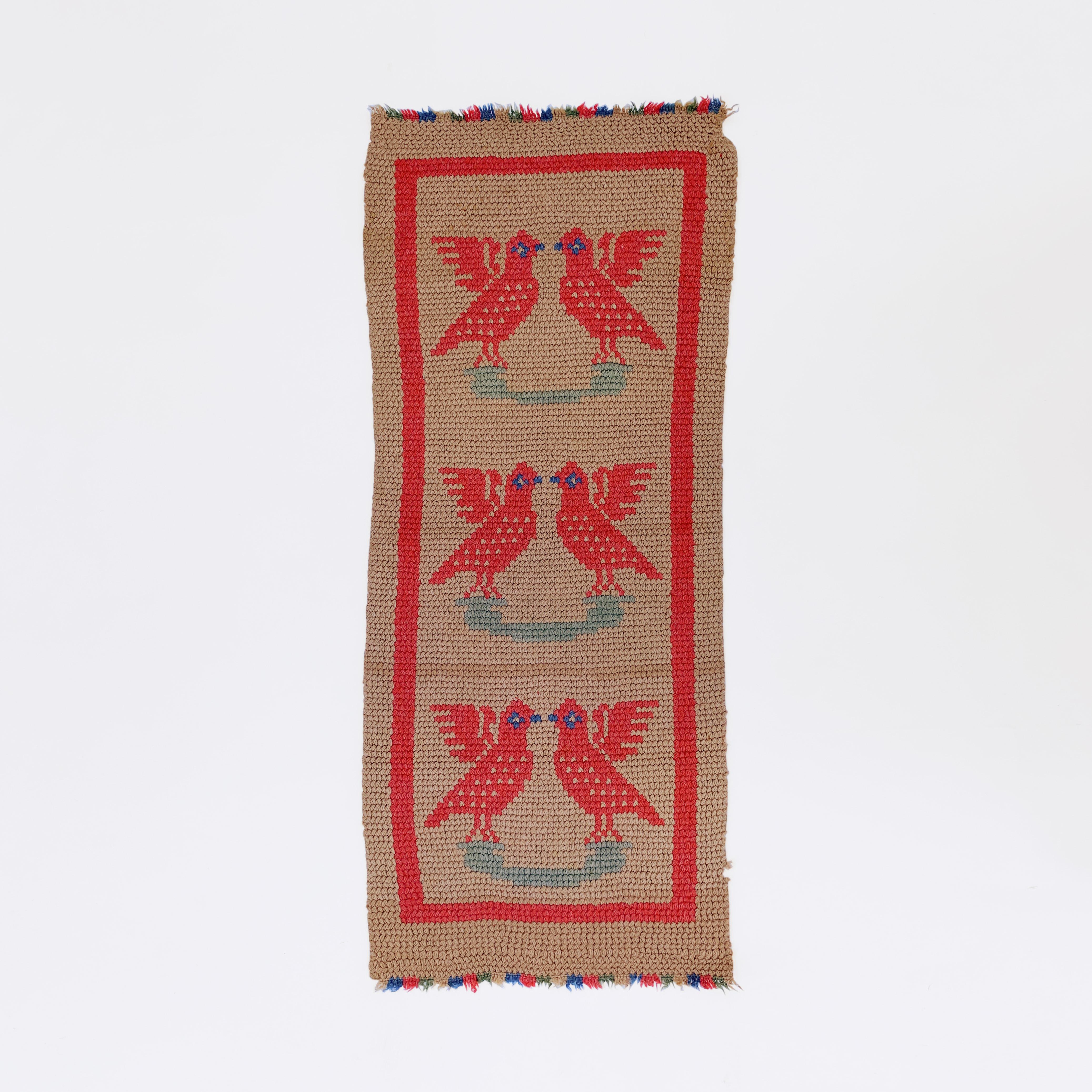 This exquisite rug, perfect for a corridor or wall decoration, comes from the Northern Greece village of Vertiskos. Hand-crocheted in a wool mix yarn, this runner features a repeating pattern of two red birds sitting on an olive branch, facing each