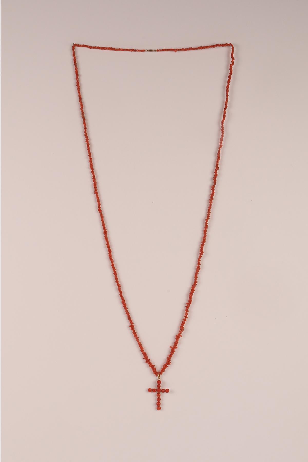 A thin and long necklace constructed from fragments of Sciacca (Sicily) coral perforated in an irregular sequence is closed by a low-carat gold clasp with an elongated barrel. A regular cross hangs from the necklace, made with a succession of