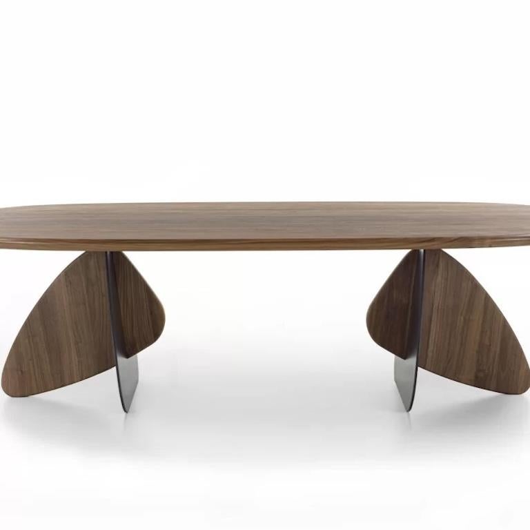 Table with a solid wood top with glued slats, with rounded corners, characterized by two legs, each composed of two volumes with soft contours, one in iron and the other in wood, meeting to create a game of intersections inspired by the shapes that