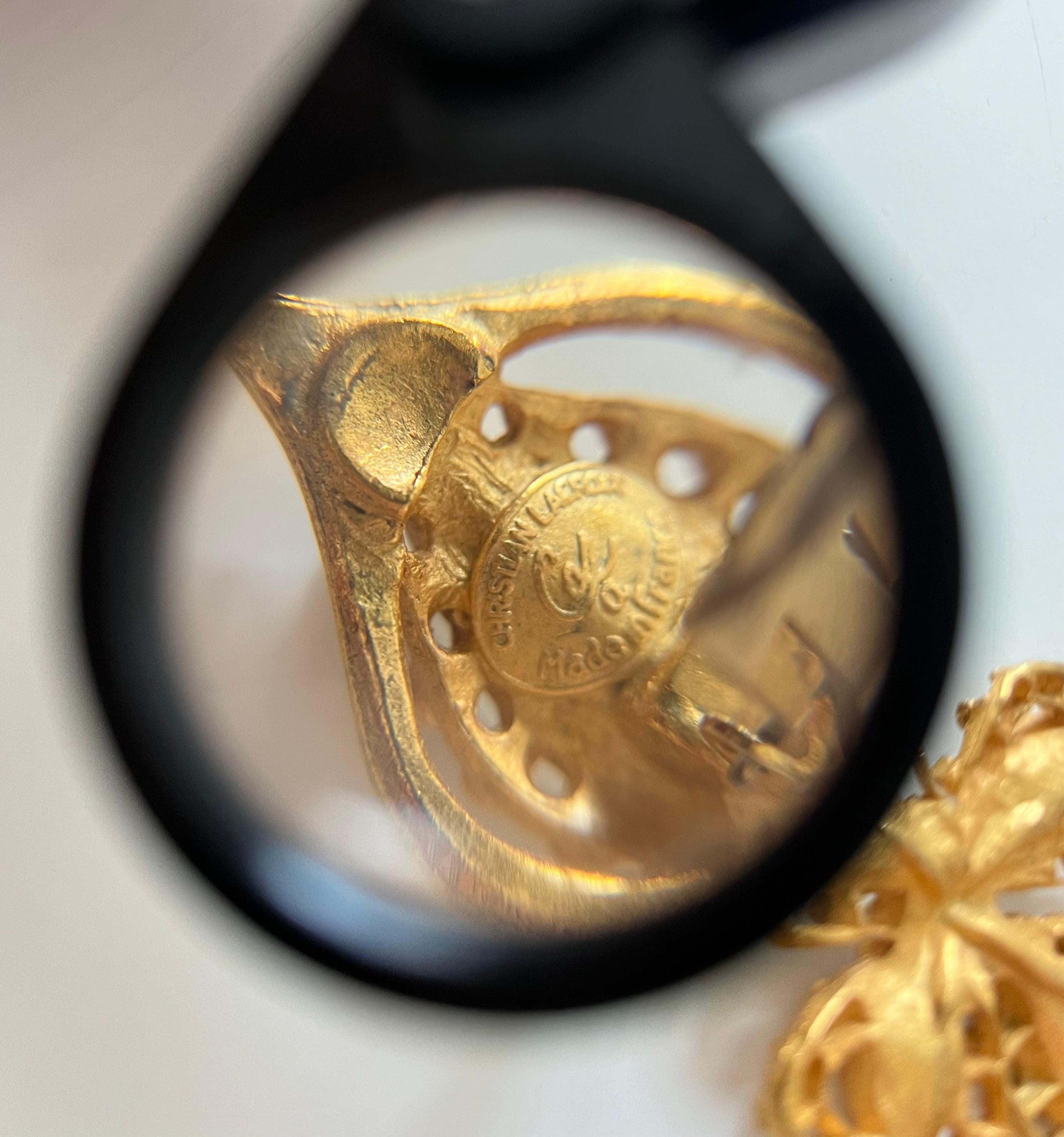 This pair of clip-on earrings is signed by Christian Lacroix (see photo). Made of gold-tone metal, this piece is in the shape of a cross. It features 4 white zircon cabochons, cut in pear and marquise shapes, on each arm of the cross. Another