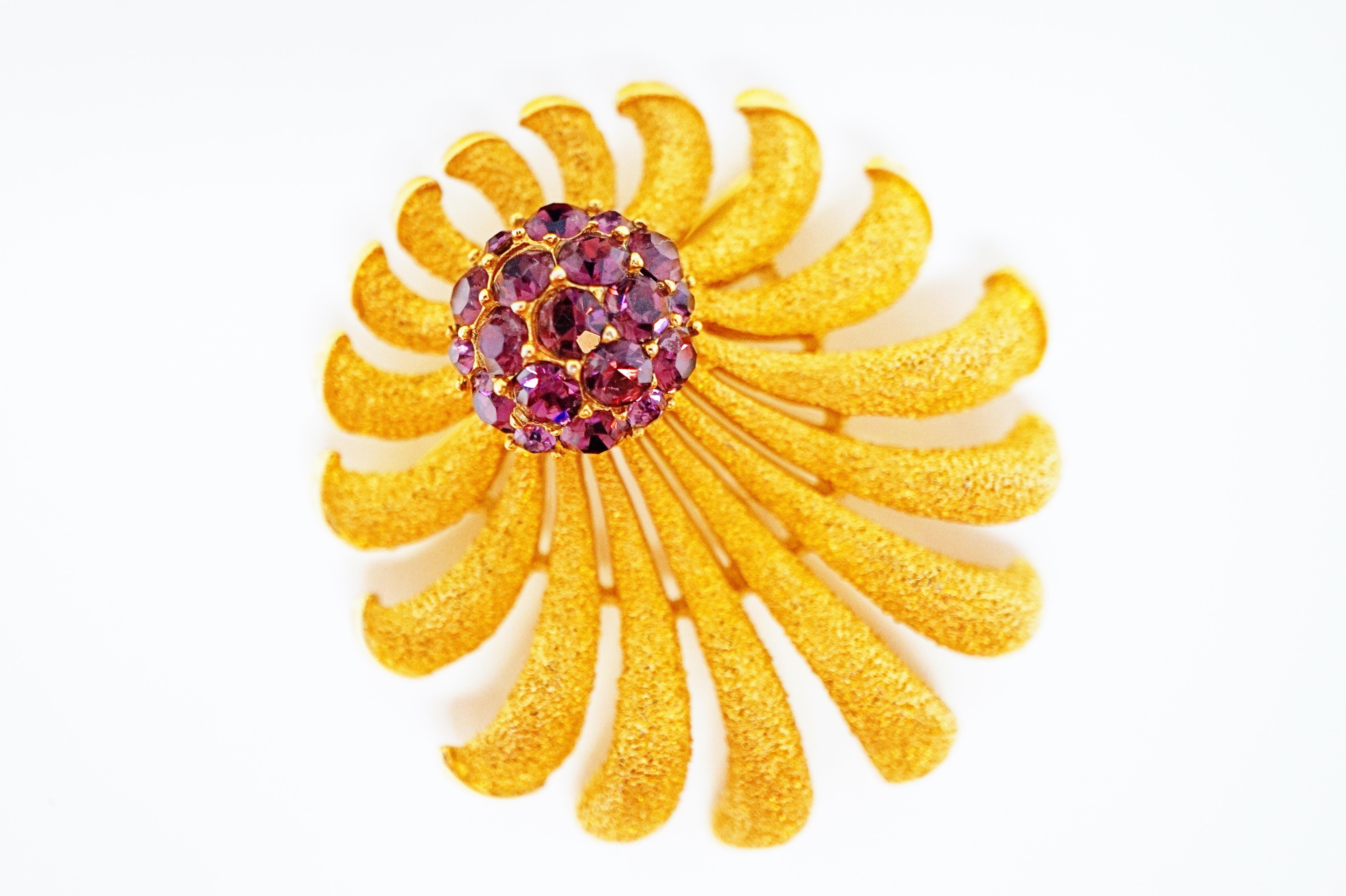 This beautiful gilded brooch by Trifari (circa 1955) marries two popular mid-century modern aesthetics - a classic sunburst design with a Brutalist-inspired texture and accented with a sparkling Amethyst-colored rhinestone cluster. A wonderful
