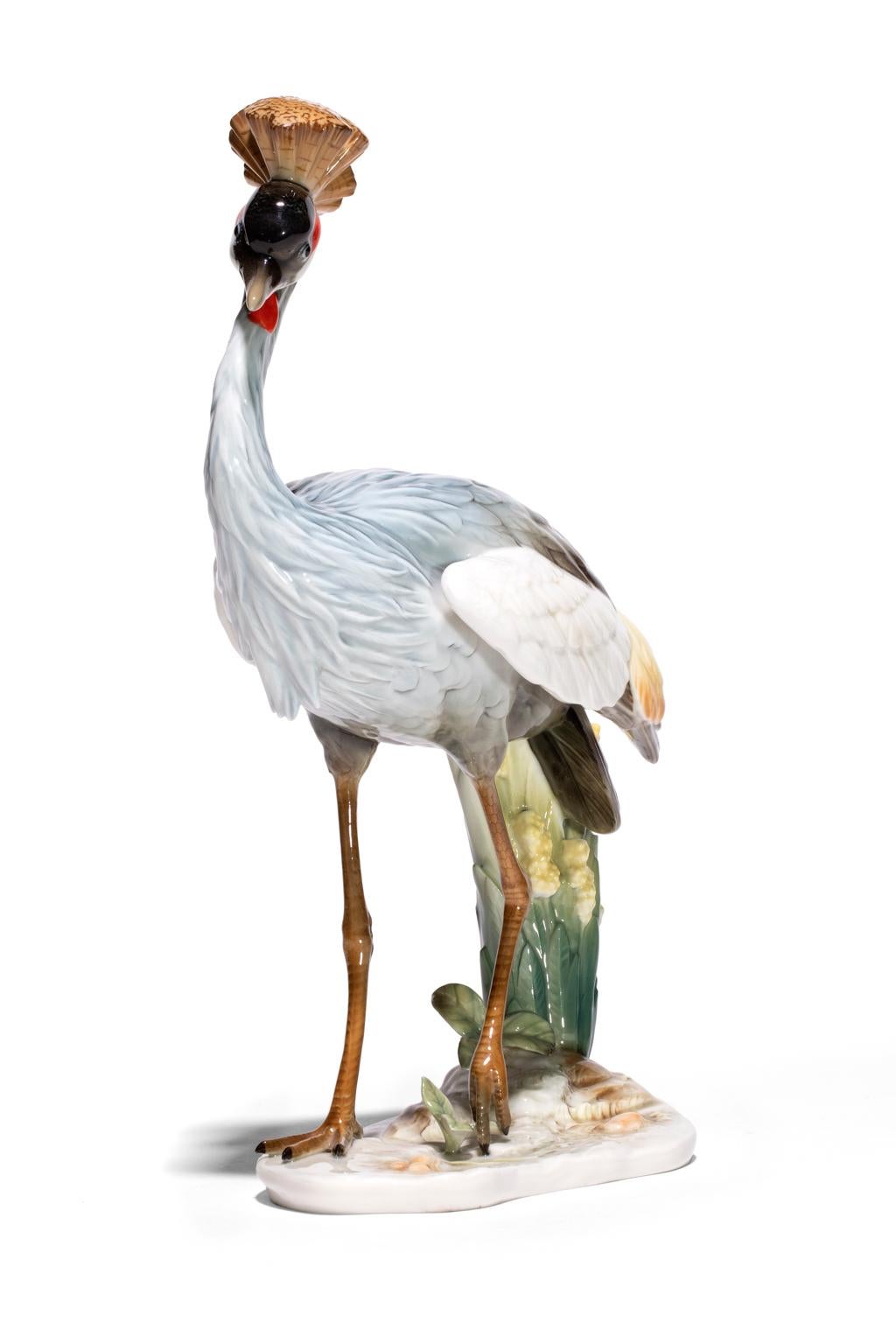 The Crowned Crane by Hutschenreuther- Selb is a richly rendered glaze painted in blue grays, maroon, pale yellow, brown and delicately rendered green foliage. In incised marks of the feathers and crown contribute to the overall precious quality of