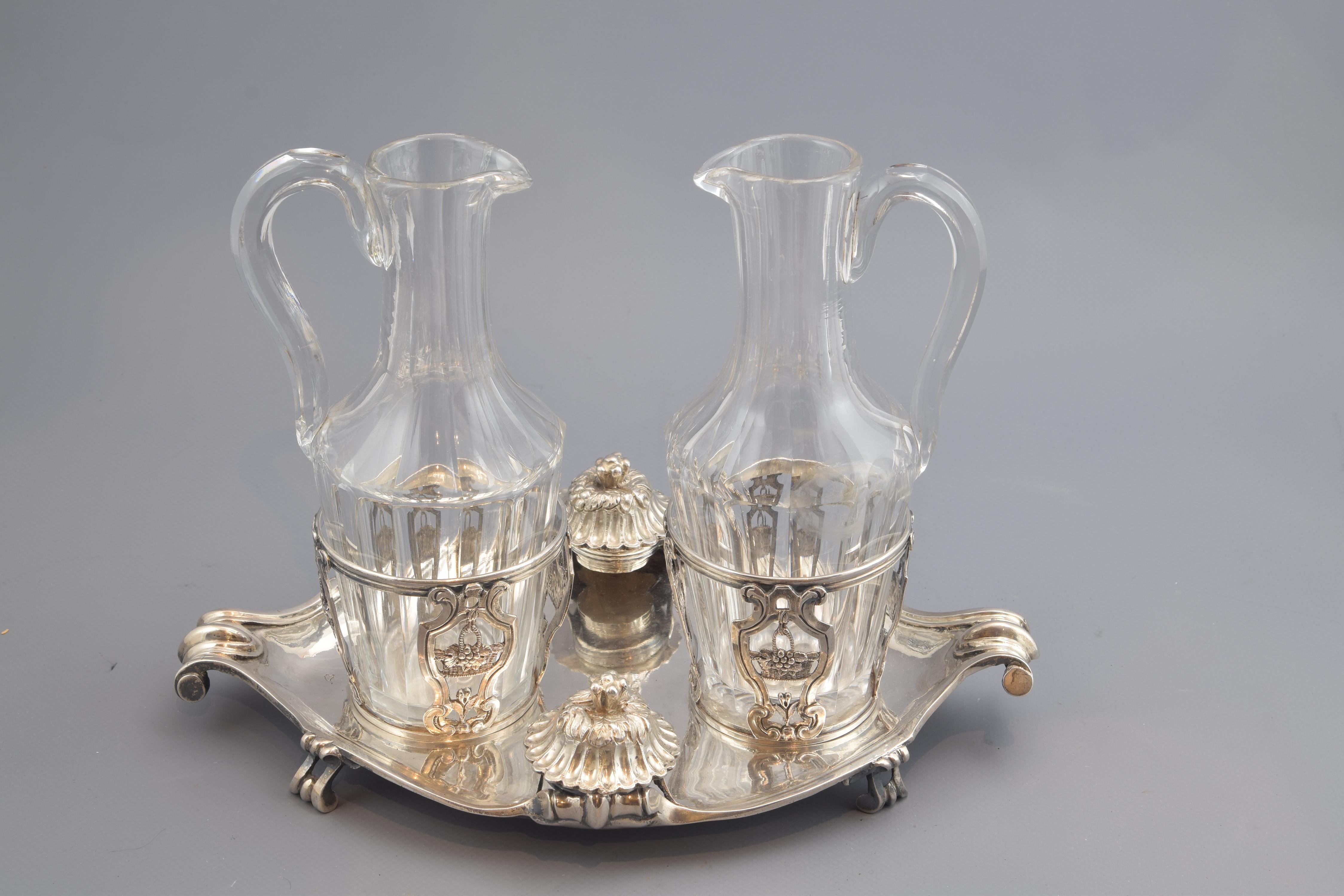 With hallmarks.
Set made in silver in its color and glass composed of two jugs with handle, elongated neck and peak, and body of circular base, with the faceted exterior, an oval tray with two supports for the jars and other two for the plugs
