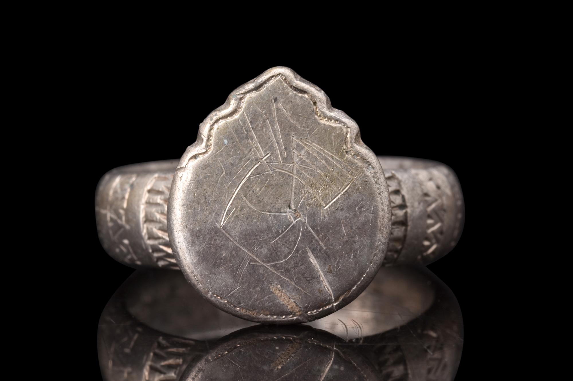 A silver ring featuring a broad, flat-section shank, providing both stability and comfort when worn. The shoulders of the ring are adorned with meticulously incised patterns. The applied shield-shaped bezel serves as the centrepiece of the ring,