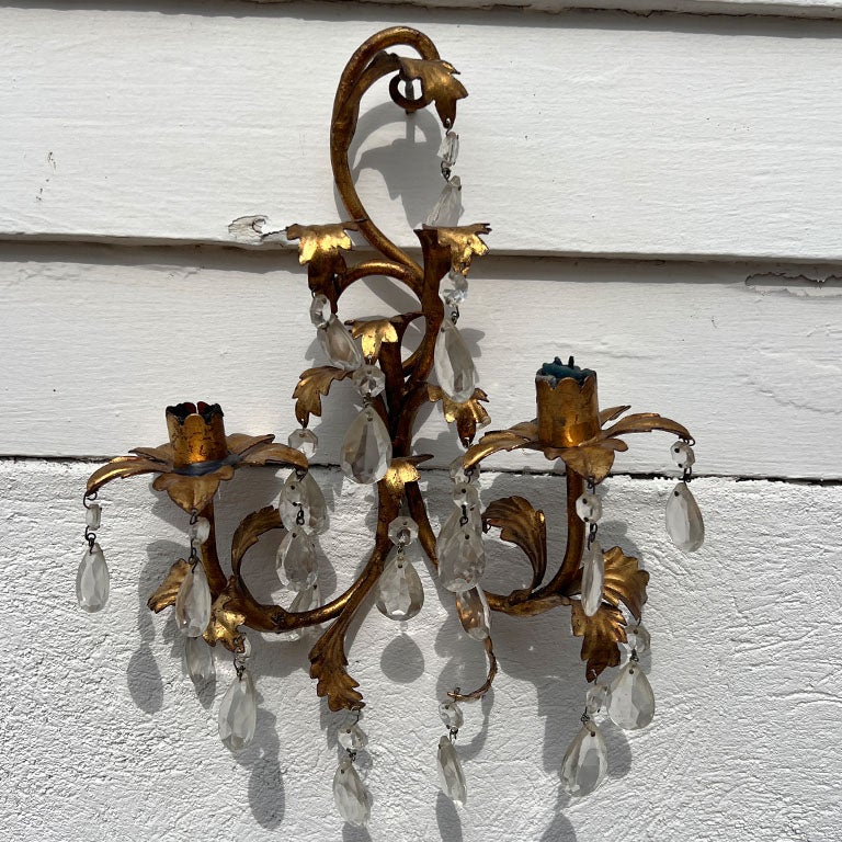 Una bellissima applique da parete in tole dorate. Questo pezzo è alto, ha due bracci e può ospitare 2 candele affusolate. È realizzato in metallo, con dettagli in tole applicati. In tutto il metallo è dipinto in oro o in foglia d'oro. Ogni braccio è