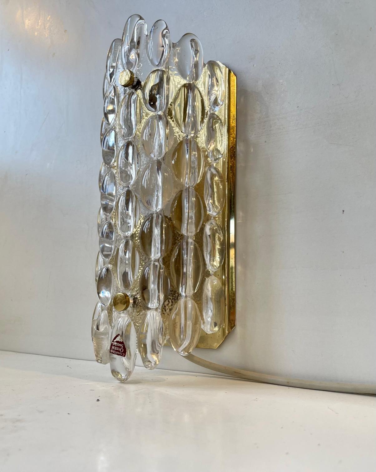 A rare crystal and gilded brass wall light designed by Carl Fagerlund and manufactured by Orrefors in Sweden during the 1950s-60s. The connected crystal drops creates a cosy, ambient and soft lighting. Measurements: H: 30 cm, W: 13 cm, Dept: 8 cm.
