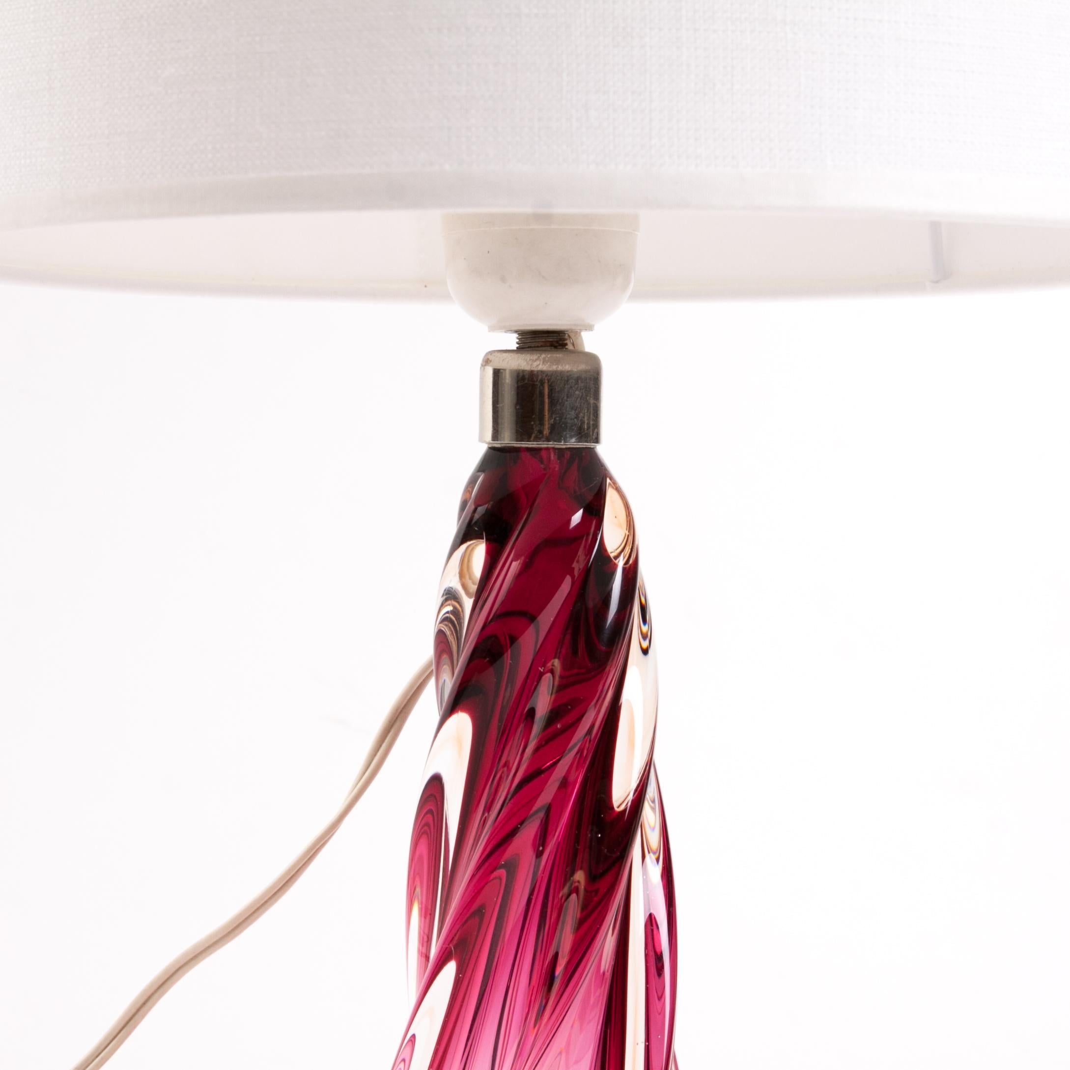 This lamp features art glass manufactured according to traditional glassworking techniques. The body, or stem of the lamp, is made of colored orange and pink glass. These colors gradually become more vivid at the base of the stem and at the top.