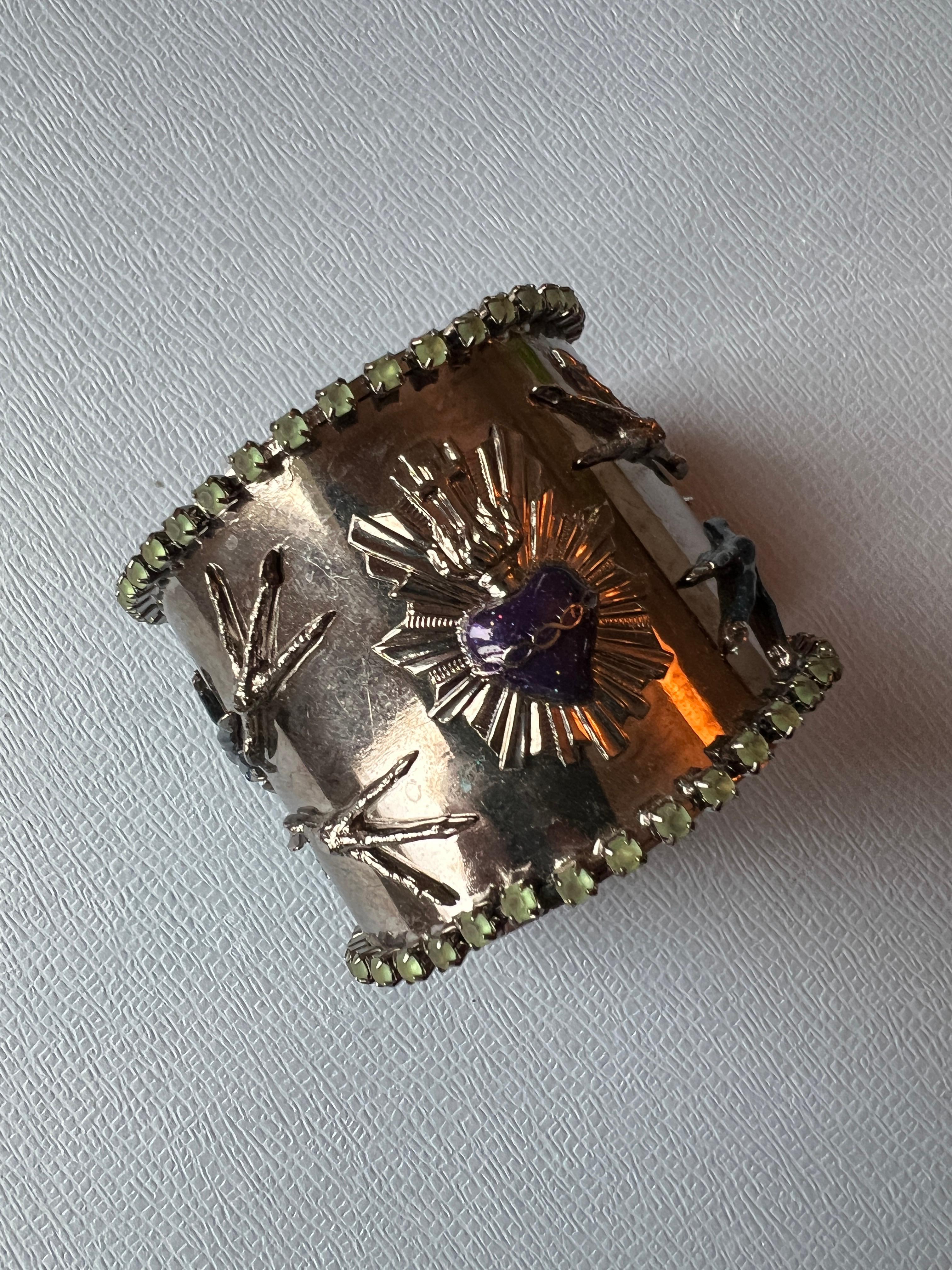 Cuff Bracelet with Sacred Heart, Purple and Blue Enamel, Green Rhinestone Border

Description:
Cuff bracelet featuring a central Sacred Heart, bird feet motifs, and initials J D at the base. The surface is hand-painted in purple and dark blue glass