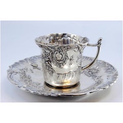 Cup and saucer in embossed silver with floral motifs surrounding a monogram card