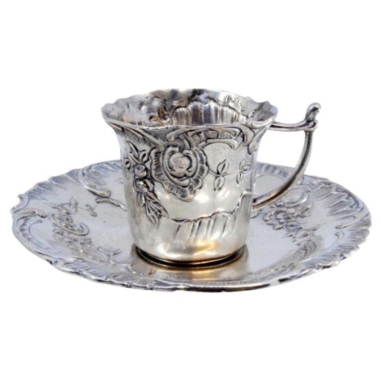 Cup and saucer in embossed silver with floral motifs surrounding a monogram card