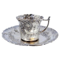 Tasse et soucoupe en argent repoussé avec motifs floraux entourant une carte monogramme.