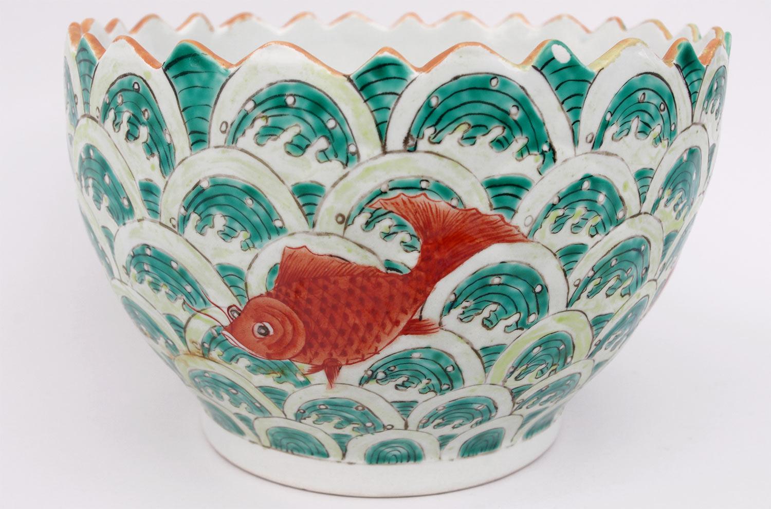 Tasse aus chinesischem Porzellan mit einem Dekor aus orangefarbenen Fischen auf grünem und weißem Hintergrund mit konzentrischen Bögen und weißen Wellen. Kante mit Wellenform.

Roter Schriftzug mit asiatischen Schriftzeichen.

Realisiertes Werk,