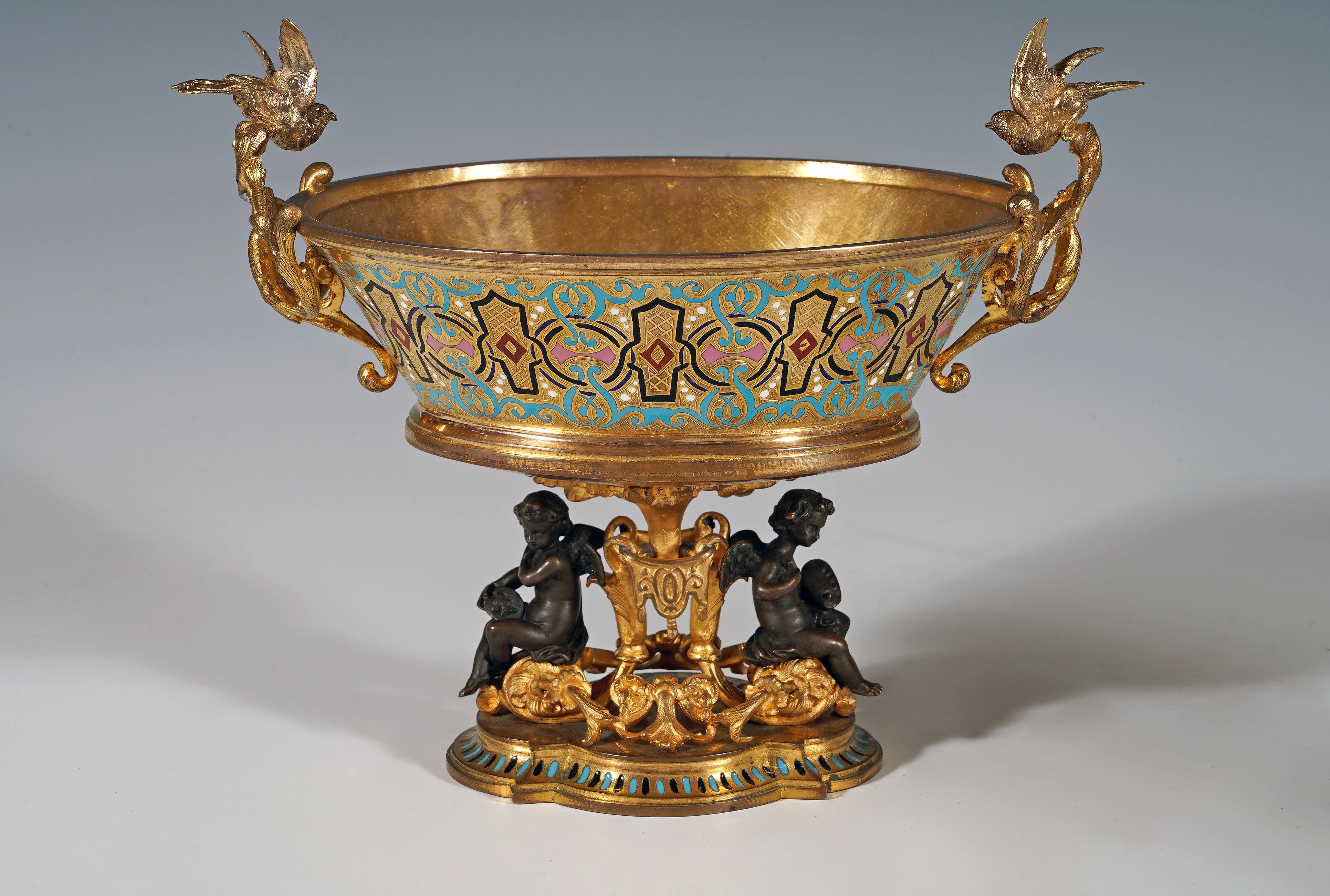Graziosa coppa di forma ovale, in bronzo dorato e smalto policromo con decorazione arabescata. I manici fogliati sono sormontati da colombe e poggia su una base polilobata sormontata da volute fogliate decorate da lambrequins su cui si appoggiano
