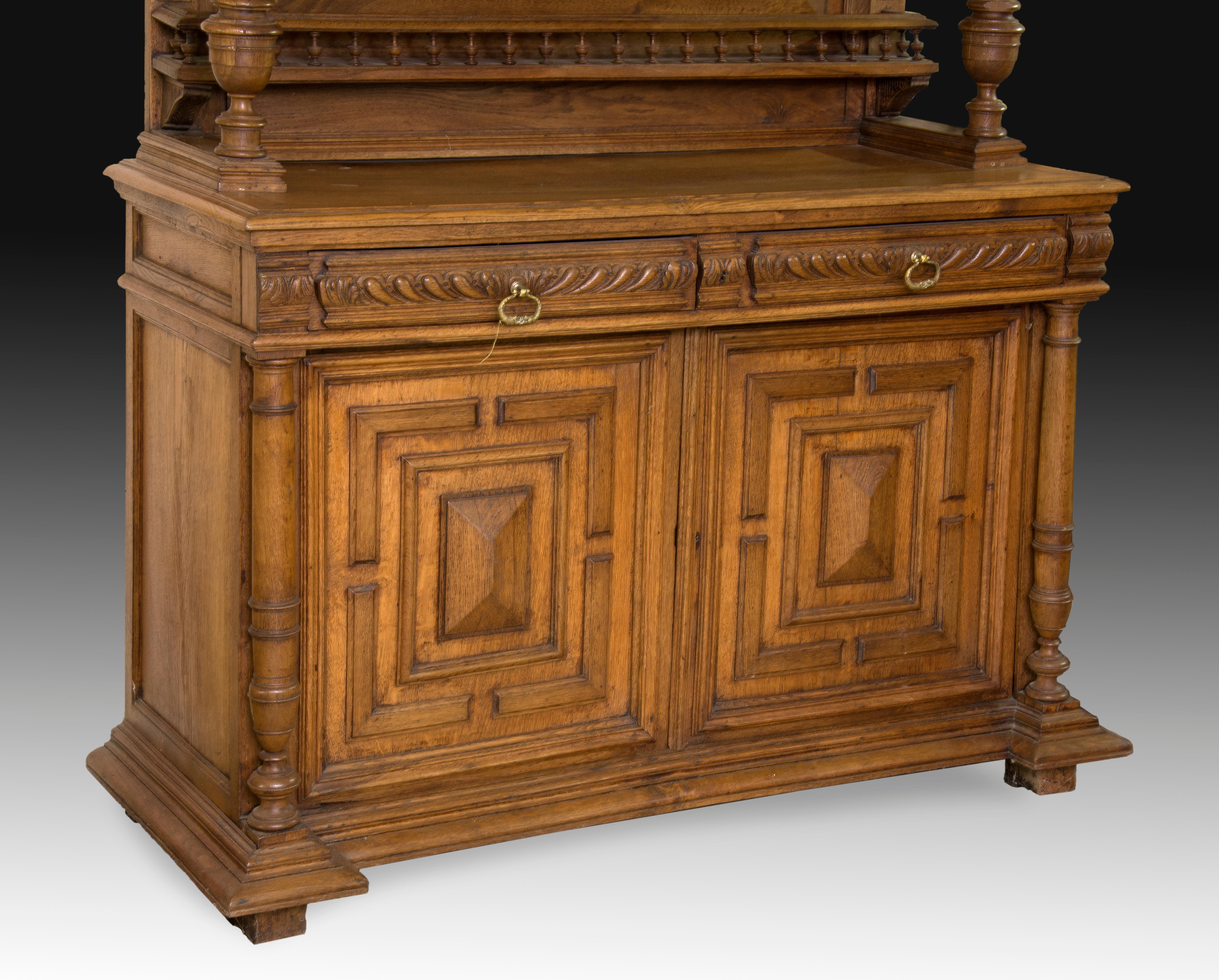Carved oakwood sideboard composed of two bodies: the upper one has a display case with shelves, and a simple decoration with columns and a powerful molding reminiscent of certain neoclassical examples; the center presents a space flanked by columns