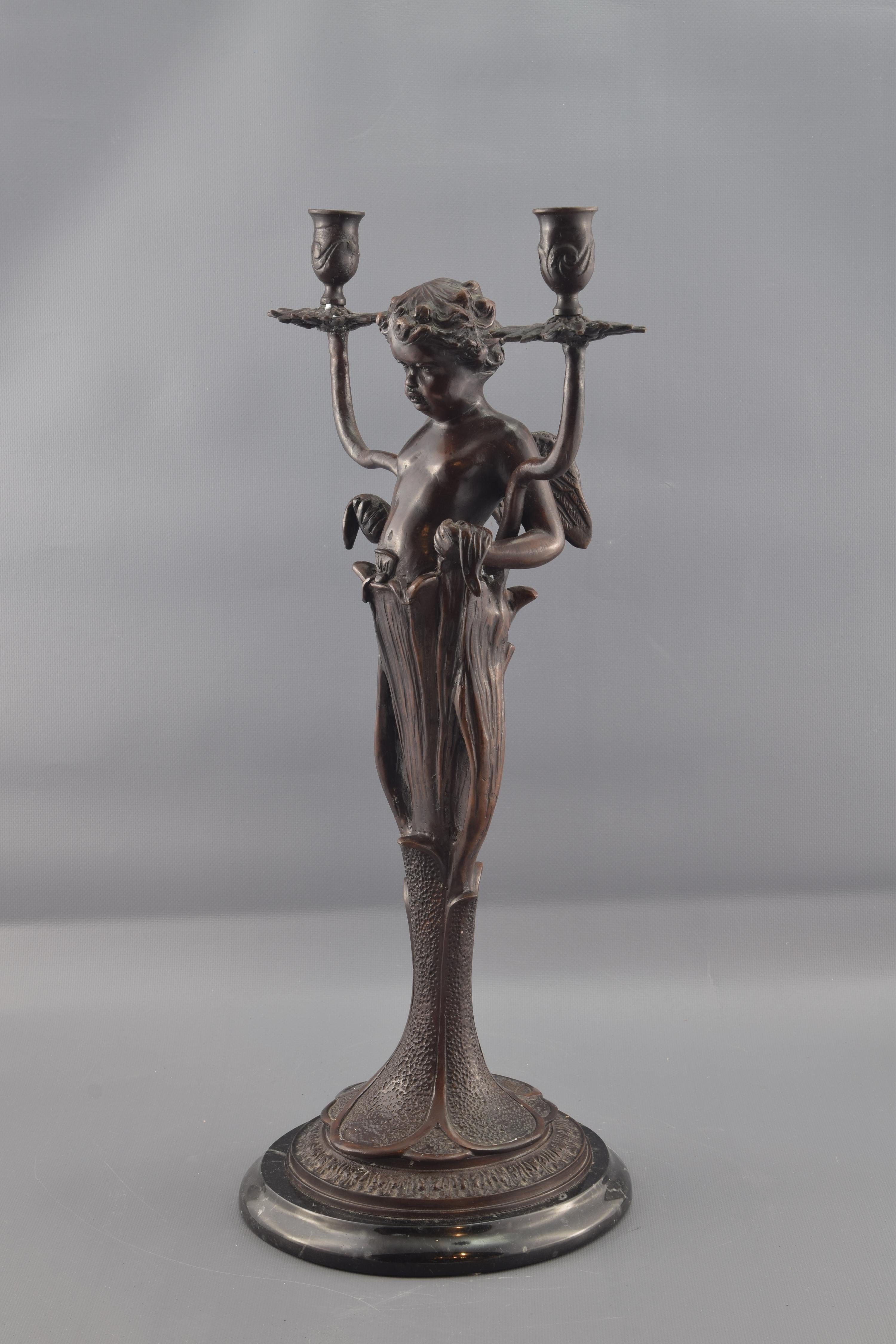 Patinated bronze. Lost wax casting. Base in marble. 
A plant stands on a circular base. Cupid emerges from it, identified thanks to its wings, which appears holding two leaves, and from its stems split the two points of light of the candelabra.