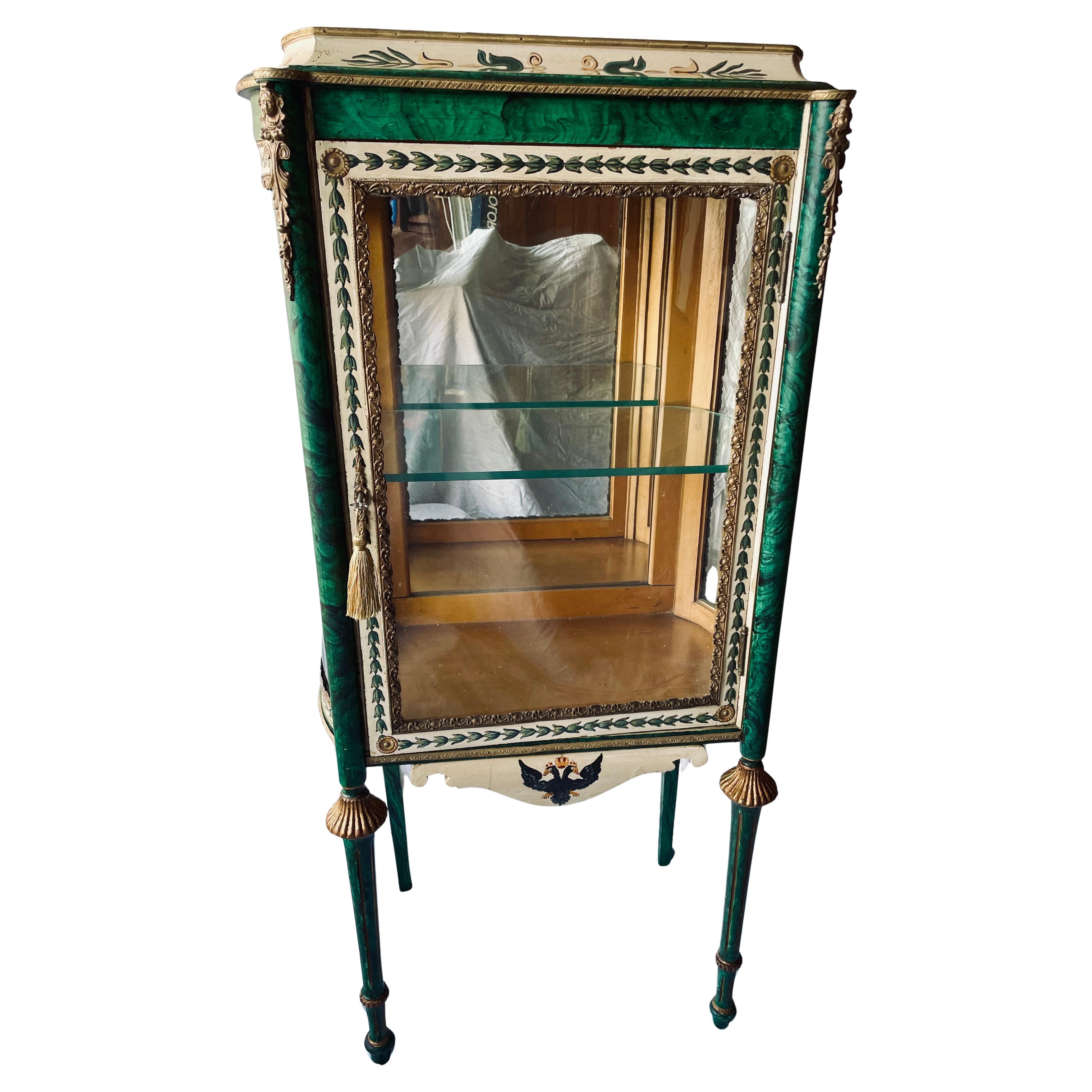 Si tratta di una splendida vetrinetta a un ripiano dipinta in stile russo con finta malachite e un'aquila a due teste sul fondo. Ci sono accenti di ispirazione francese intorno alla porta e ai lati in vetro curvato. Gli angoli superiori mostrano