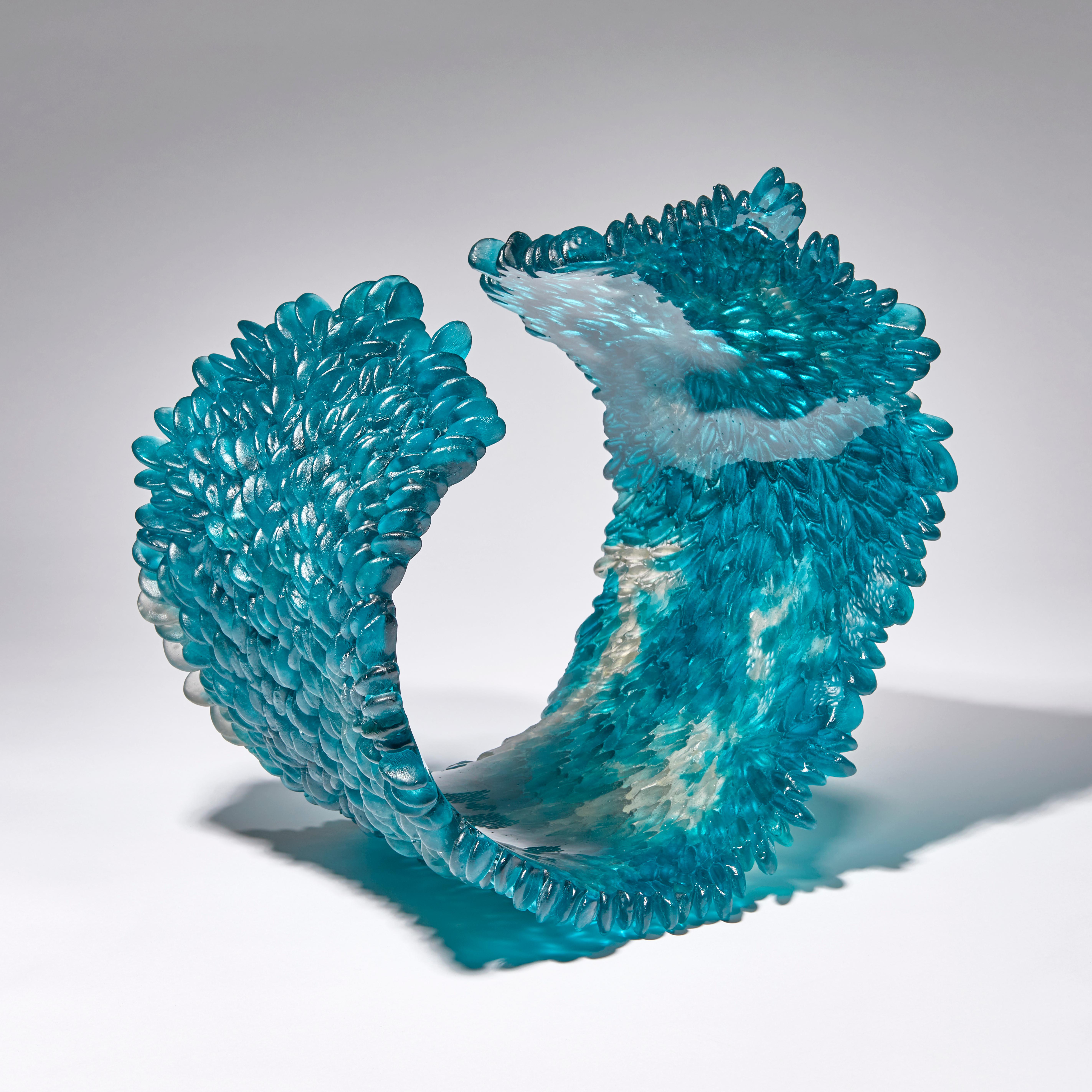Curled over V is a unique textured cast glass sculpture in teal blue or deep aquamarine and grey by the British artist Nina Casson McGarva.

Casson McGarva firstly casts her glass in a flat mould where she introduces all of the beautifully