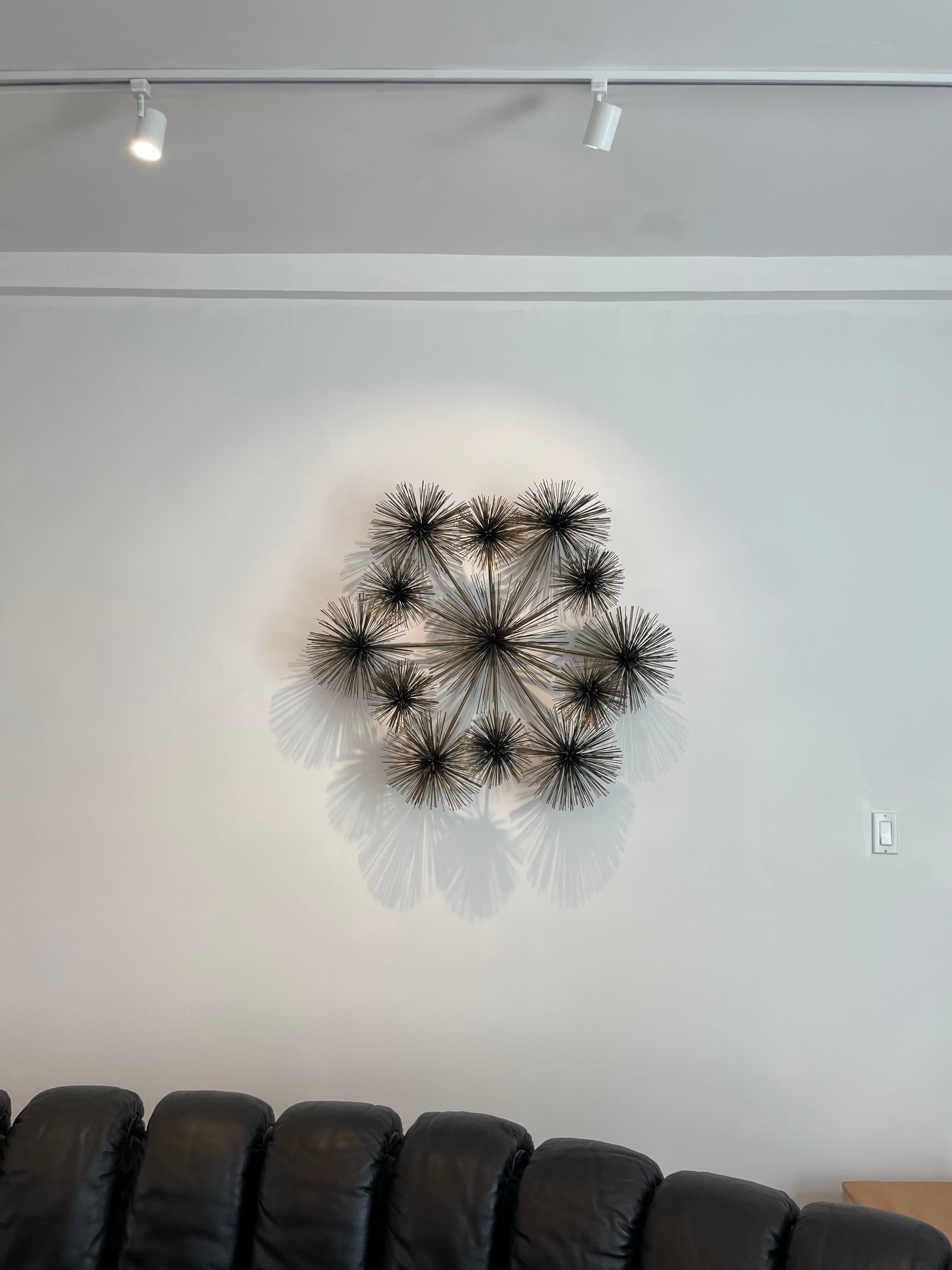 'Dandelion' wall sculpture in brass by the artisan brand Curtis Jeré of the Artisan House collective. It has 13 dandelion flower figures that also look like sea urchin shaped 'star bursts' are that are welded in place in symmetrical formation: 12