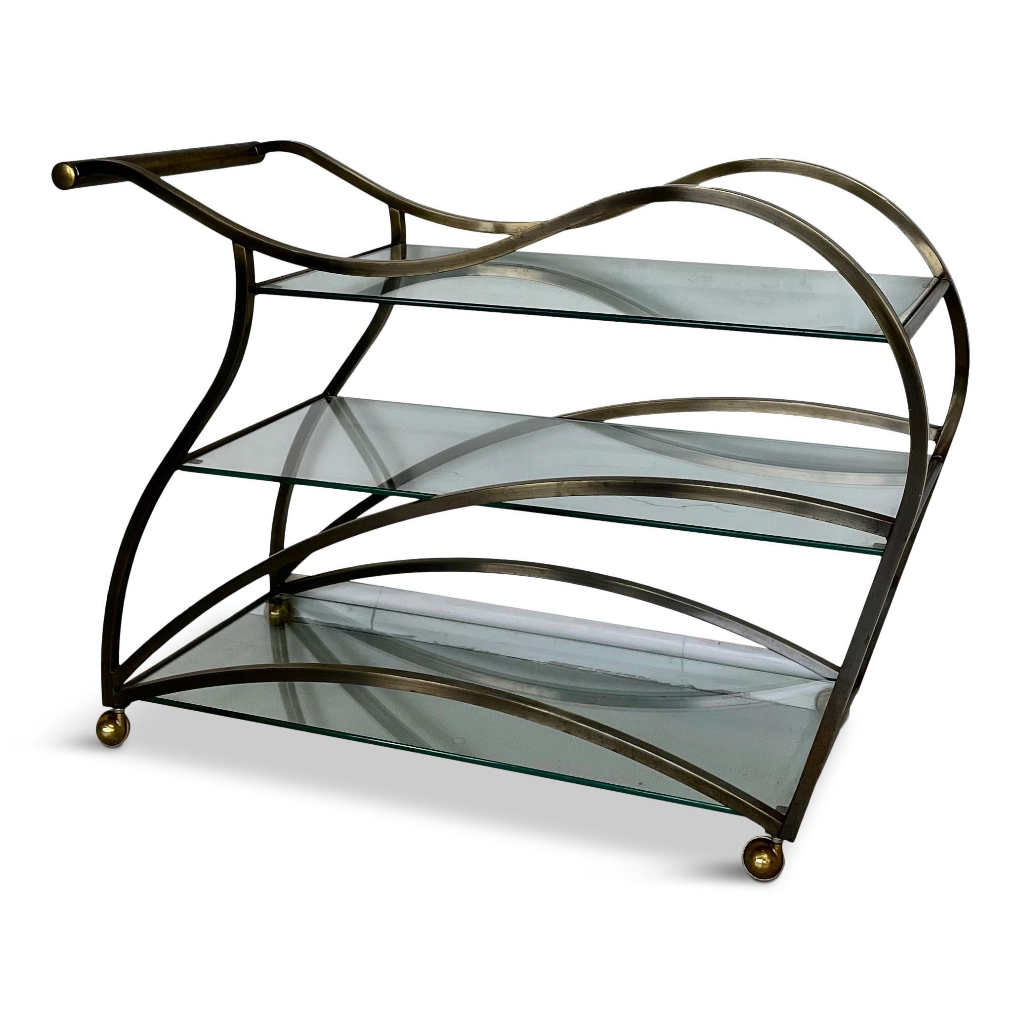 Sexy and curvaceous sculptural bar or tea cart manufactured by high quality furniture maker, Design Institute of America. This rolling cart has a metal frame with an antiqued brass finish and three, 3/8 inch thick, clear glass shelves. Its large