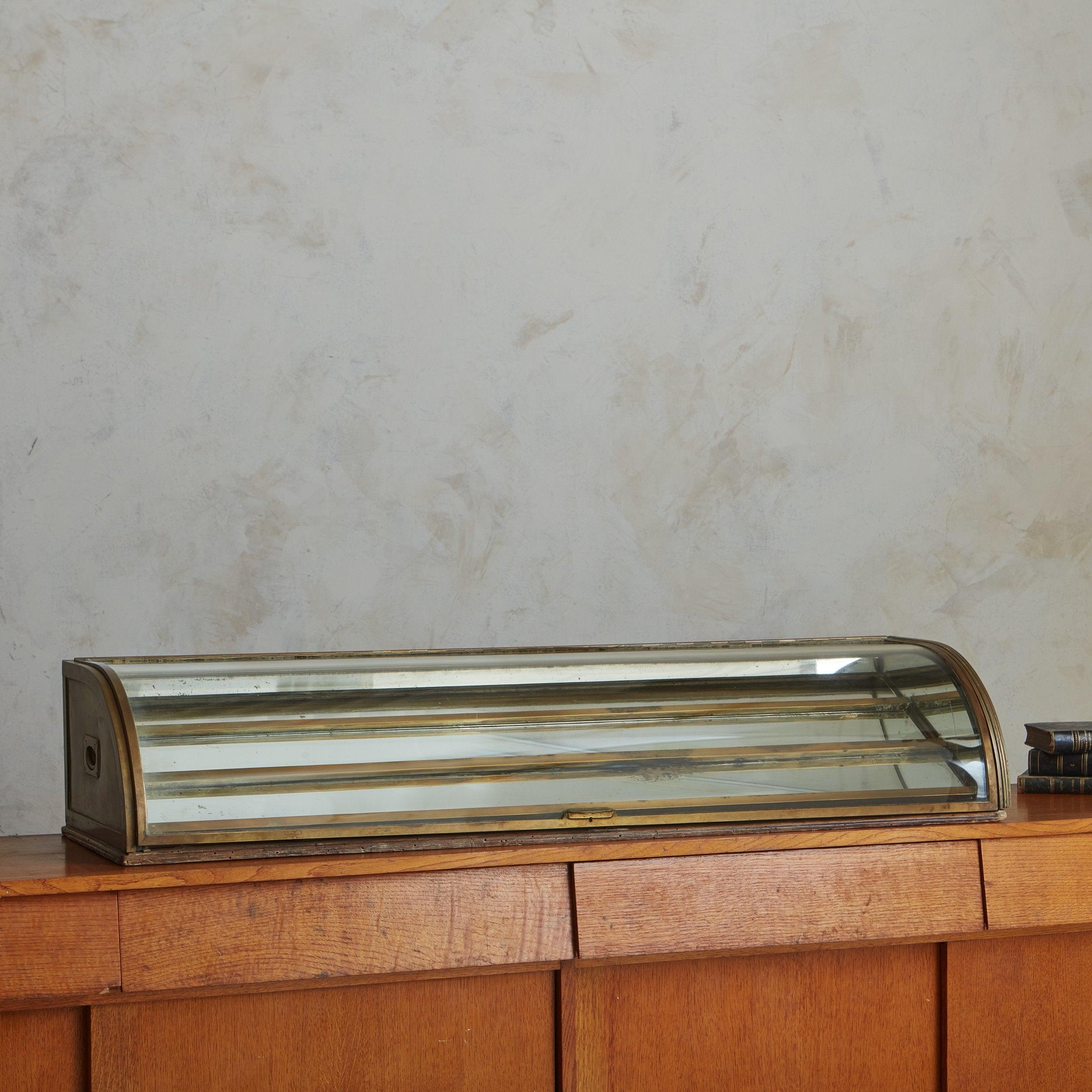 A 1920s French showcase display used on a shop or drugstore countertop to display candies and other boutique inventory. This elegant piece has a patinated brass frame with two handles and a wooden backing. A curved glass lid opens outwards to reveal