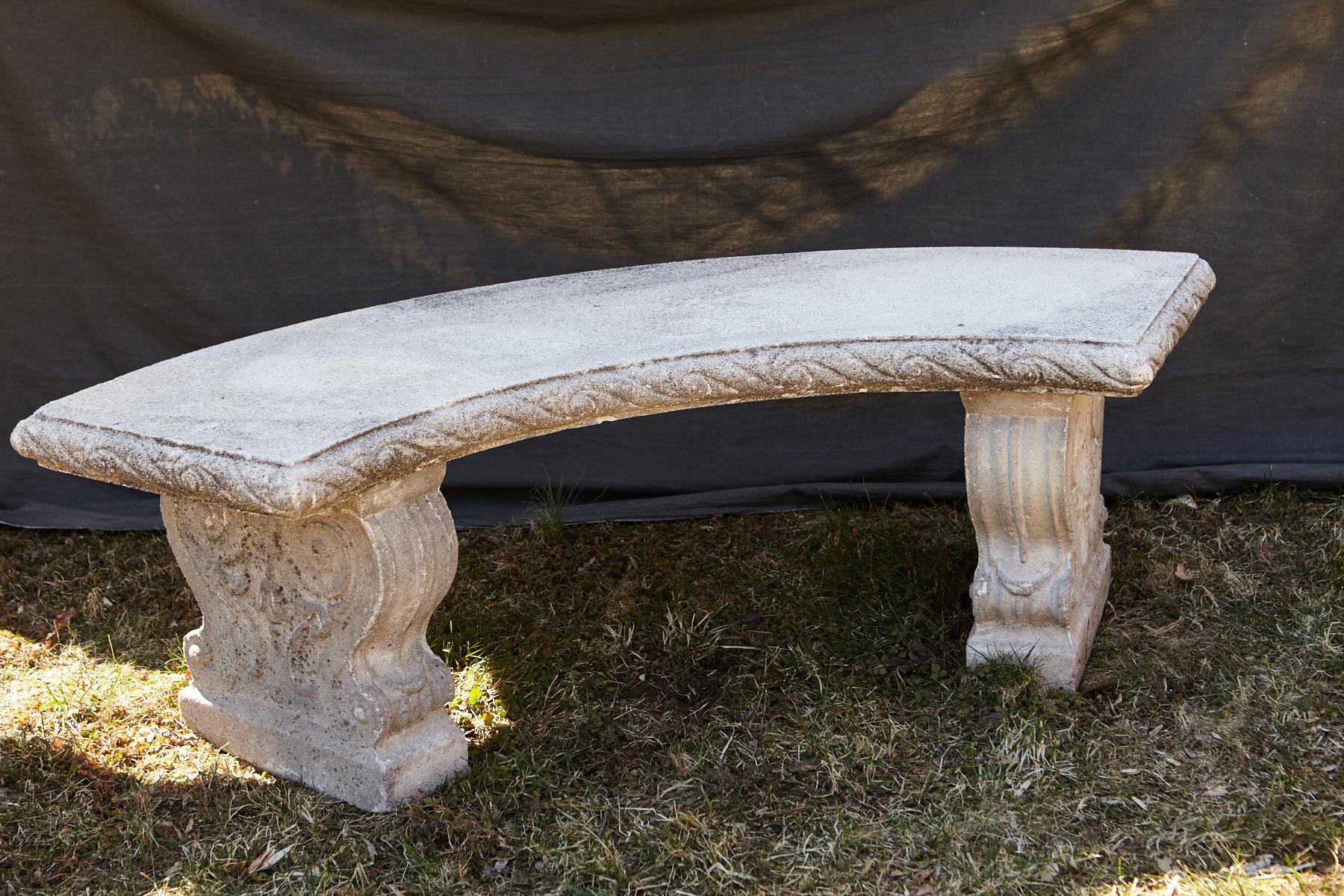 Curved cast stone bench with an ornamental carved band of alternating concave and convex sections around the flat seat, reminiscent of waves, mounted on carved legs with carved double sided scrolled ornaments and leaves.
The bench has a very nice