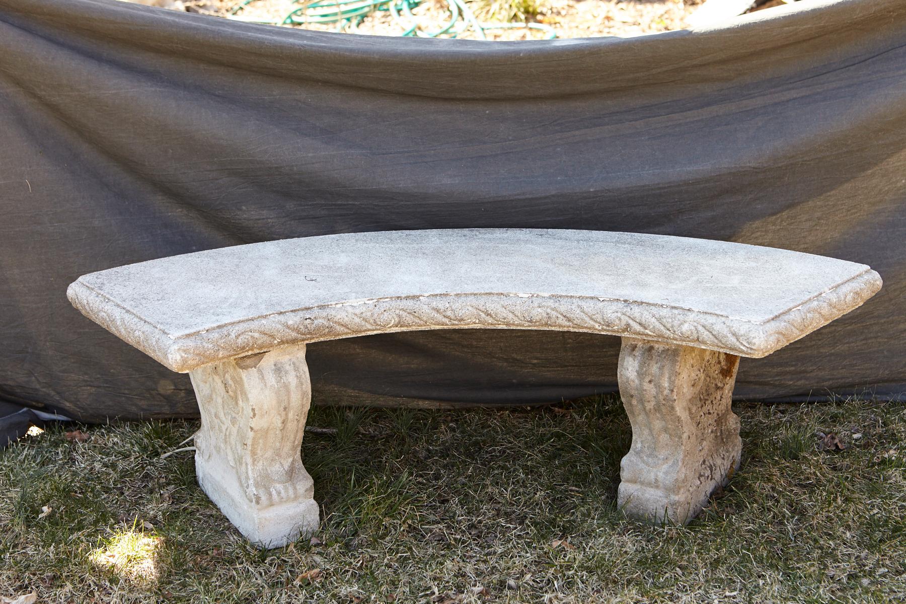 Curved cast stone bench with an ornamental carved band of alternating concave and convex sections around the flat seat, reminiscent of waves, mounted on carved legs with carved double sided scrolled ornaments and leaves.
The bench has a very nice