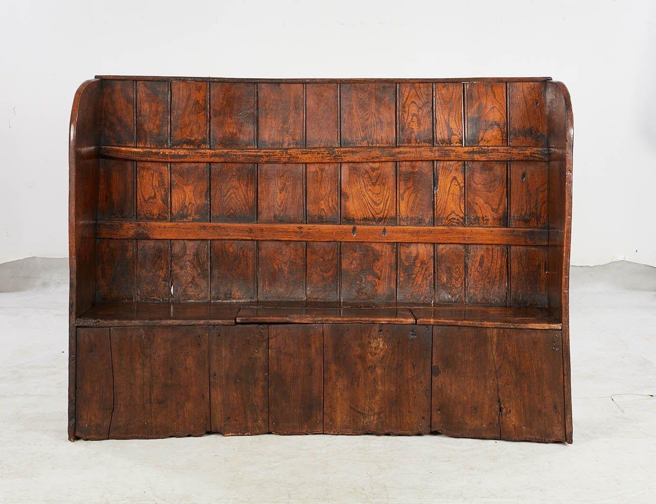 A curved high back settle with winged sides and lateral strapping bars and featuring a lift top bench seat for storage. In elm with stunning color and figuring. Would make a practical and attractive seat in a mudroom or a useful room divider between