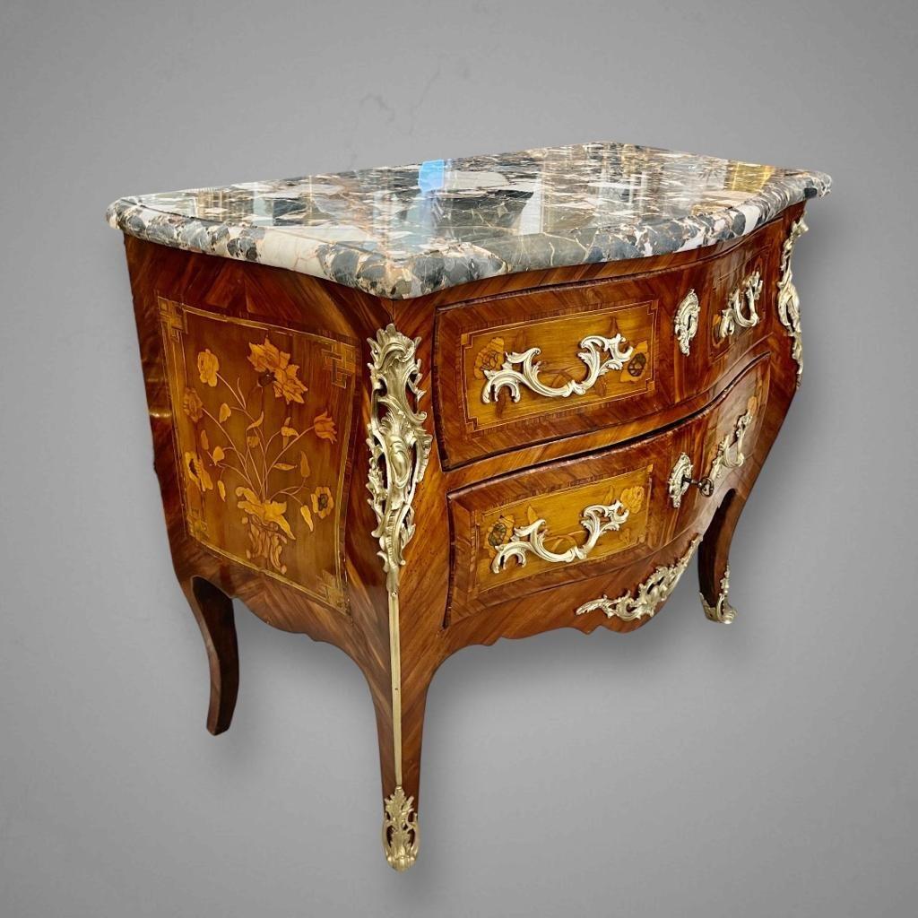 We present you this wonderful 18th-century chest of drawers in the transitional style of Louis XV features a curved design and delicate floral marquetry patterns. It comes with two front drawers, showcasing finely crafted floral marquetry that adds