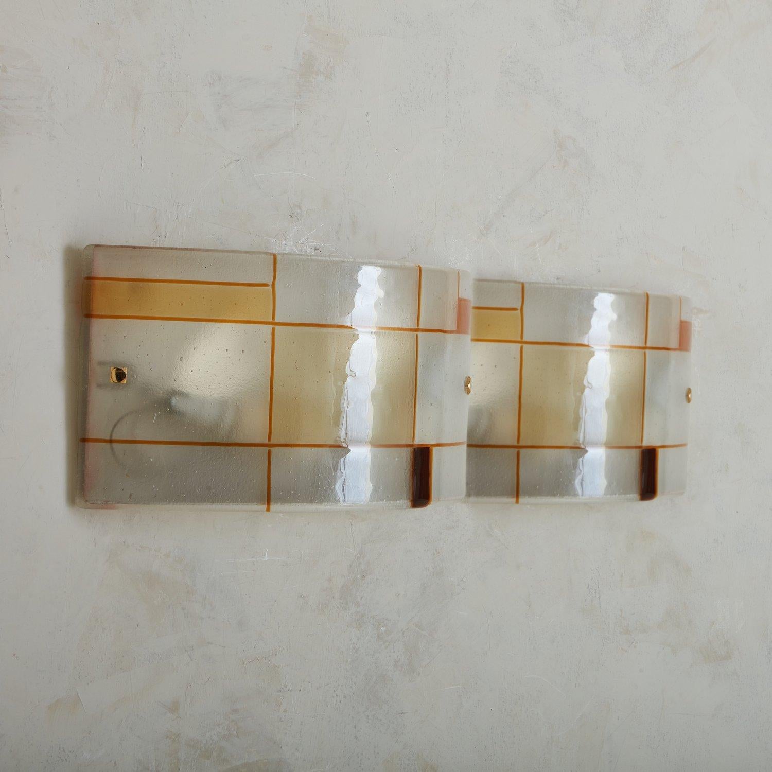 A 1960s hand blown Murano glass sconce featuring a curved shade with a Mondrian-inspired geometric design with orange and amber hues. The shade sits in a demilune cream metal base with squiggle cutout details. This sconce has brass hardware and can