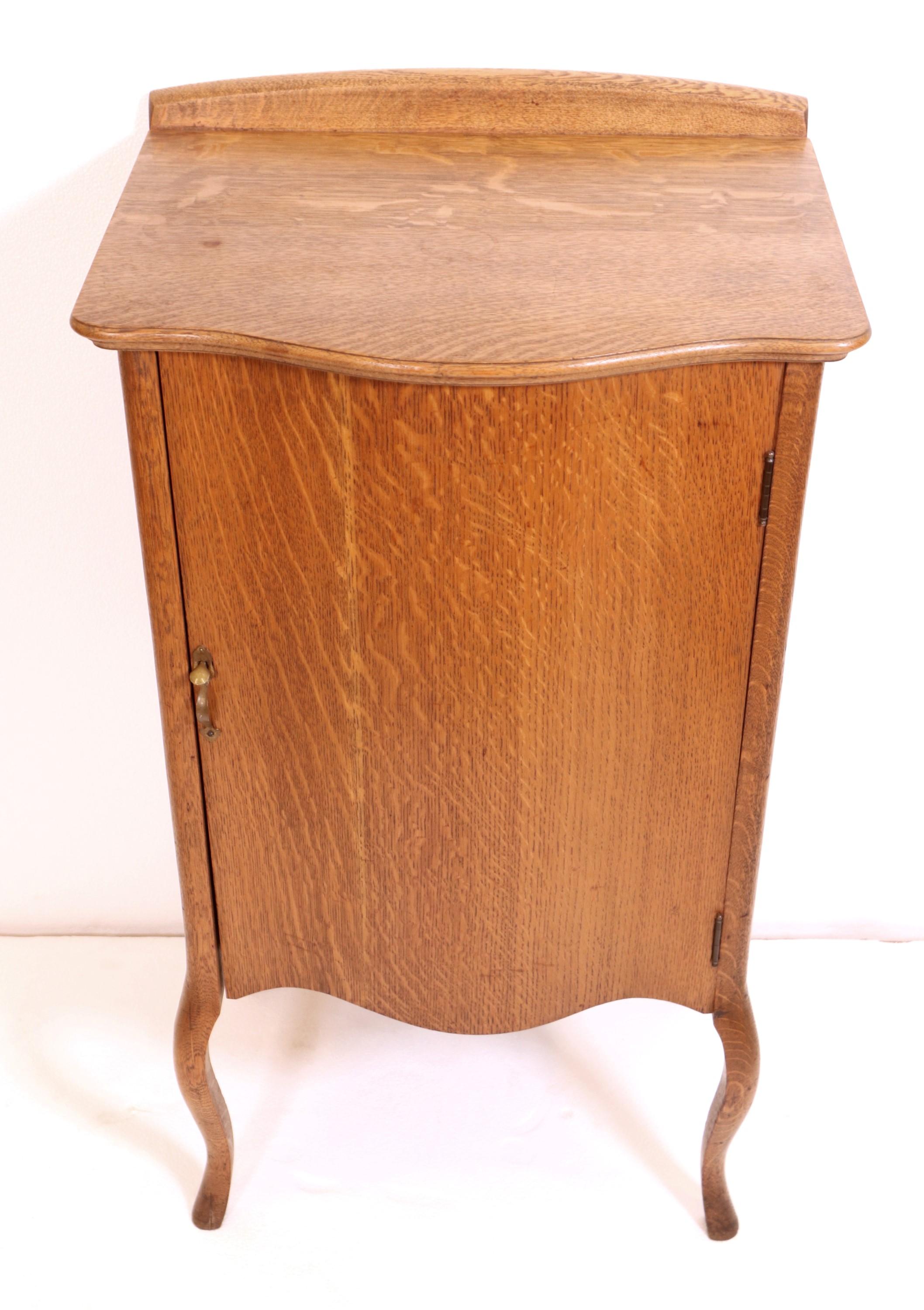 Antique oak sheet music cabinet with curved front and cabriole legs. It has an oak veneer and the cabinet door has a brass handle. There are five music sheet shelves inside the cabinet. This can be seen at our 2420 Broadway location on the upper