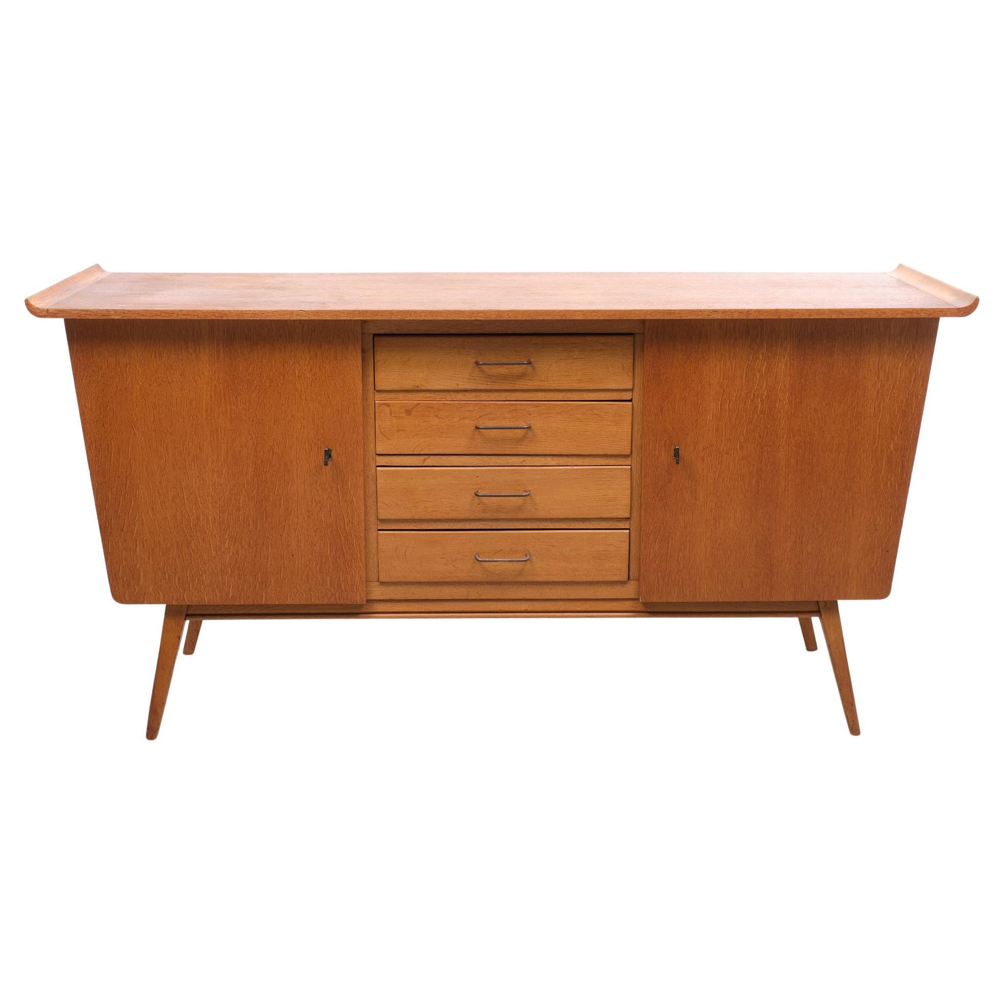 So typical 1950s This Light Oak sideboard. Very nice curved ends on the top.
4 drawers in the middle. Brass handles.Two doors left and right. Comes with the 
original keys.