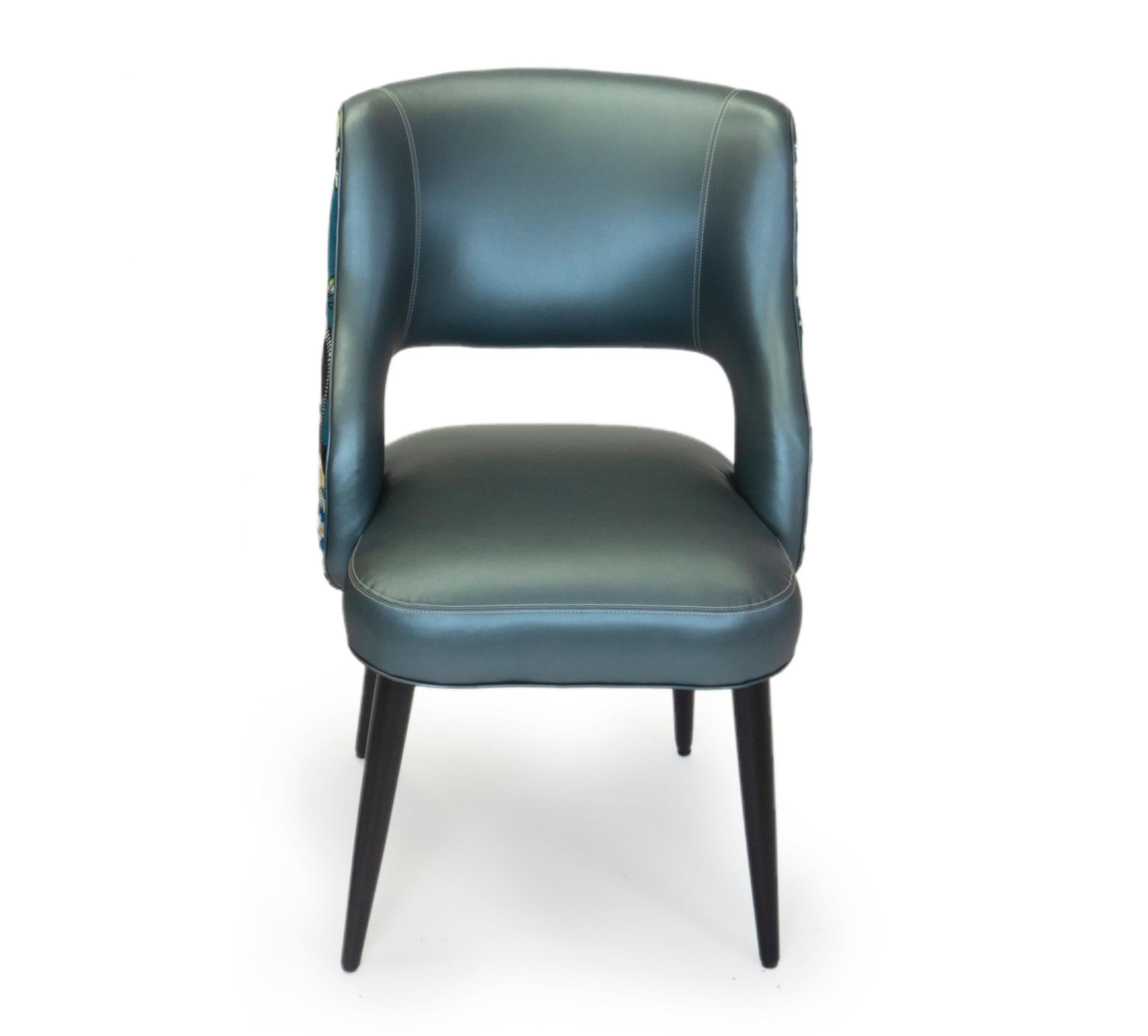Ce nouveau design de chaise de salle à manger est confortable et moderne, avec un ton décontracté. Les chaises présentées ont une finition en noyer et sont recouvertes d'un vinyle bleu. Le dos est un tissu Christian Lacroix présentant un motif