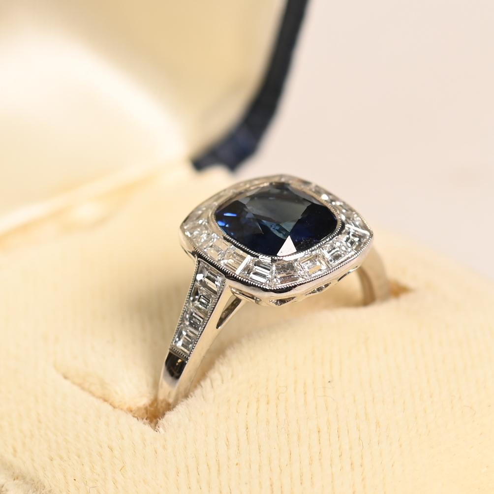 This exquisite cushion cut sapphire with baguette set diamond ring showcases a striking approx. 1.50-carat cushion-cut natural sapphire, radiating a deep, velvety hue. Framing the sapphire is a meticulously arranged halo of baguette-cut diamonds,