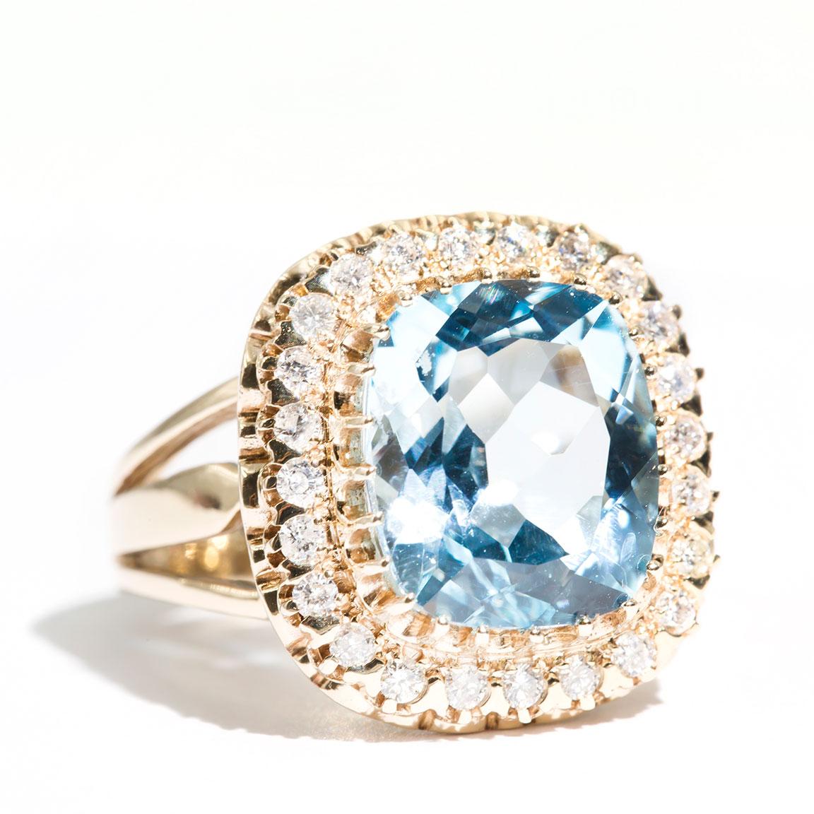 Crafted in 9 carat yellow gold, this unique vintage inspired cocktail ring features a lovely bright blue cushion cut Topaz encompassed by a halo of sparking round brilliant cut diamonds. We have named this vintage splendour The Elizabeth Ring. The