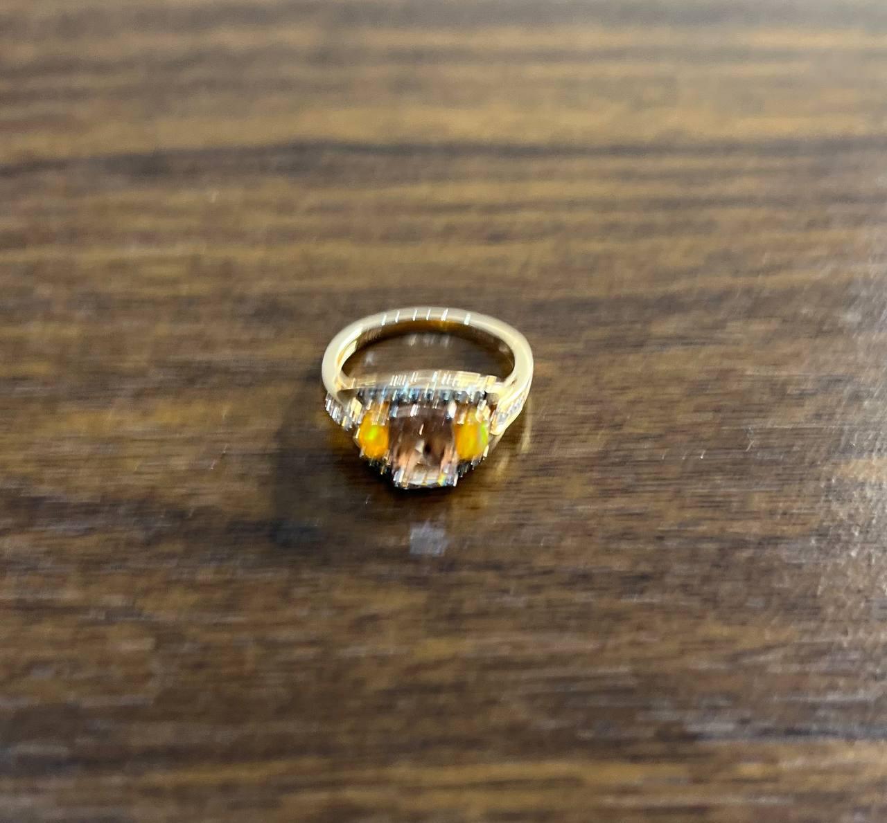 Elegant Three-Stone Ring. This exquisite ring features a central elongated cushion shape Morganite , flanked by two orange opals. The setting is intricate, with detailed metalwork surrounding the diamonds. The band is smooth and polished, providing